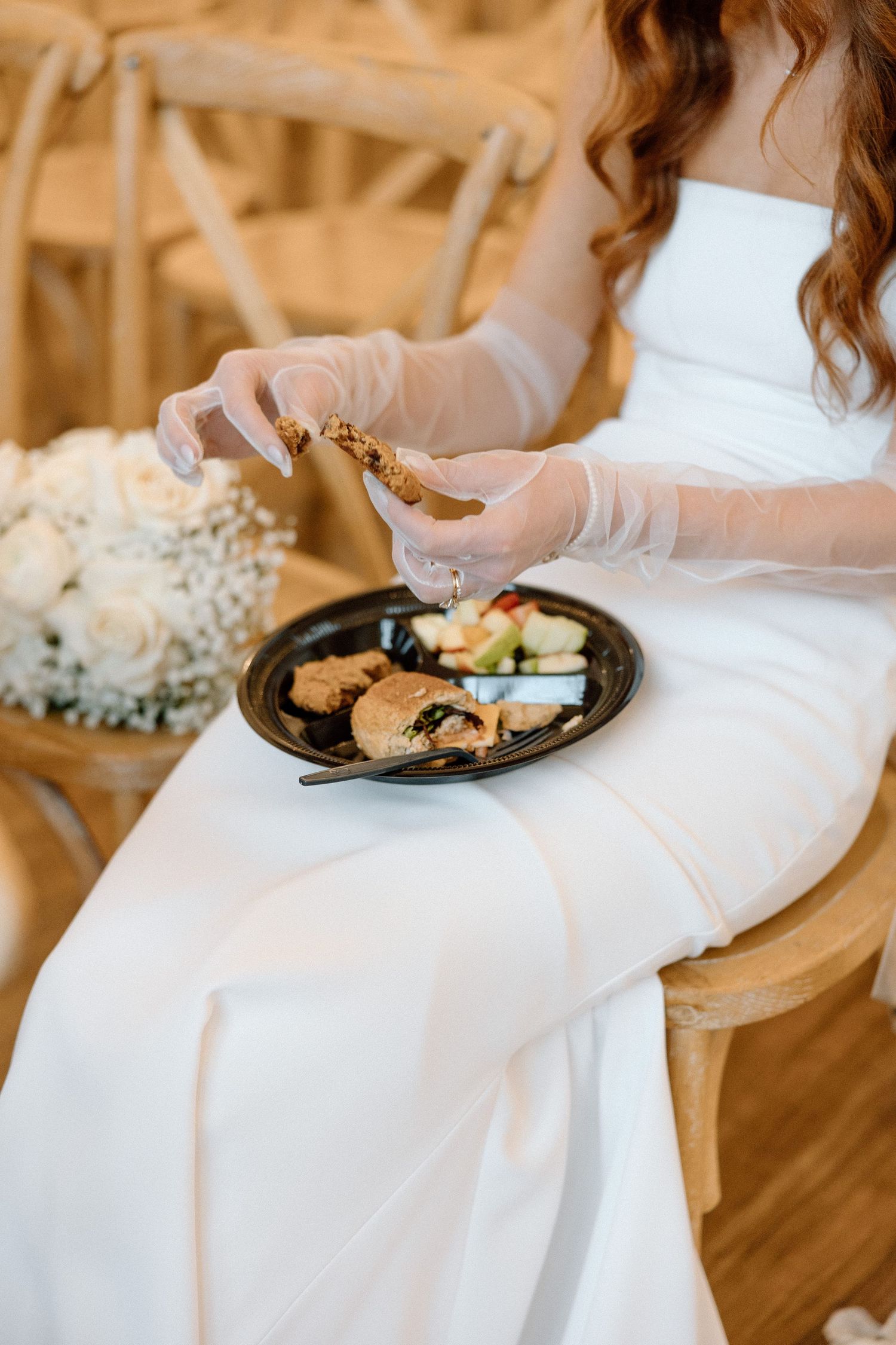 A person in a flowing white dress sits holding a plate of food at a formal event.