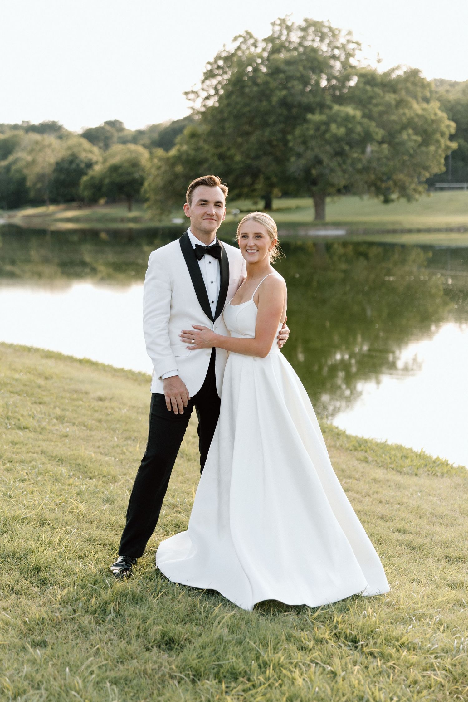 A couple in wedding attire poses together on a grassy slope near a serene lake during golden hour.