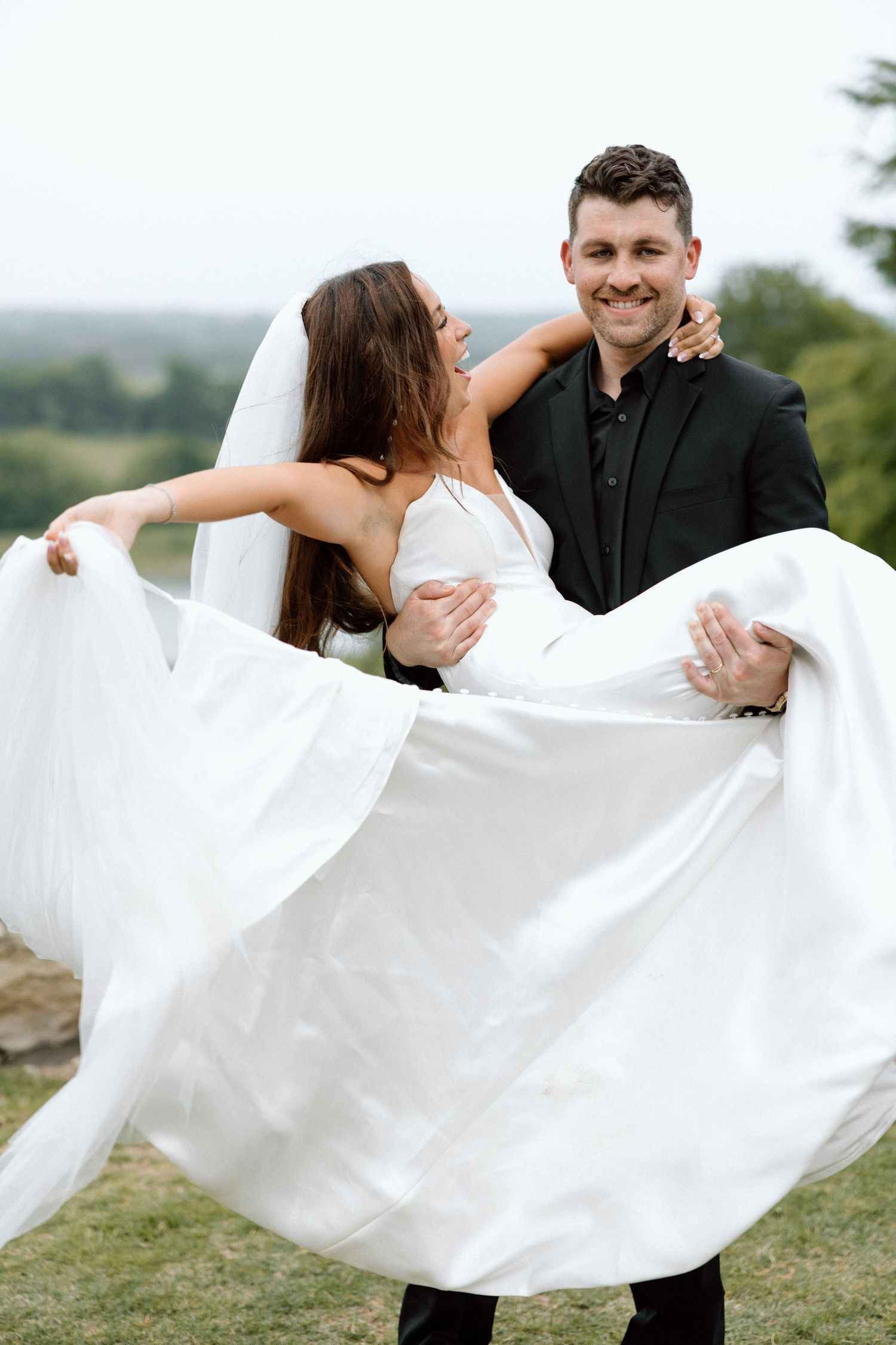 A romantic wedding couple embraces outdoors as the groom carries the bride in her flowing white dress on a hilltop.
