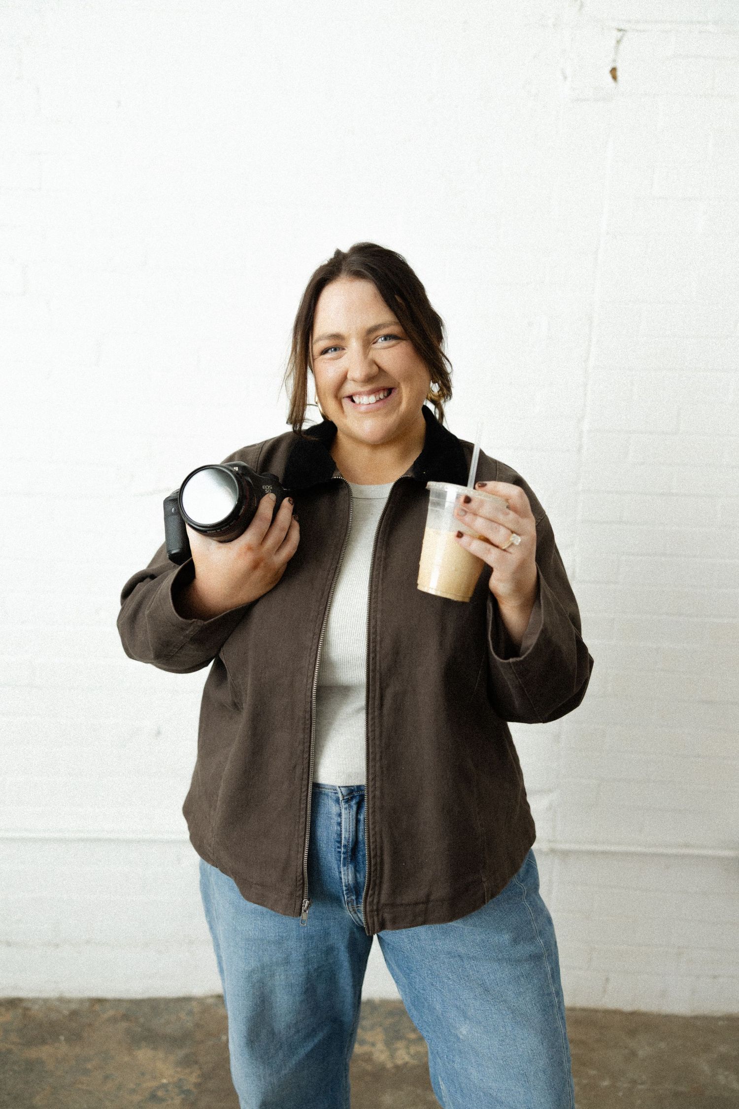 A person in a brown jacket and jeans poses playfully with a coffee cup against a white wall.