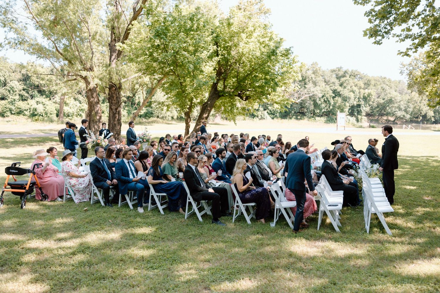 Large group of wedding guests seated in white chairs on a grassy lawn under shady trees during an outdoor ceremony.