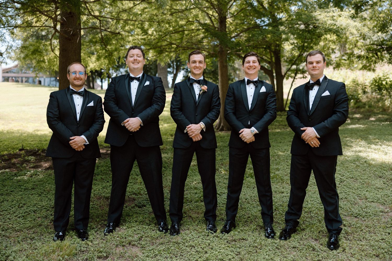 A group in formal black tuxedos poses together outdoors against a backdrop of green trees and sunlit grass.