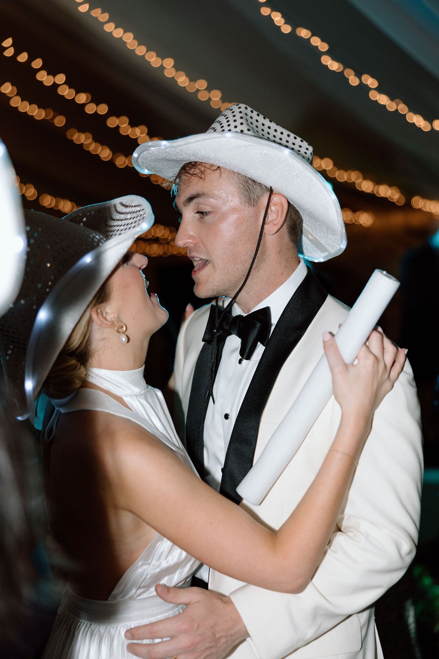 A couple in matching white formal attire dance together under warm string lights at an evening celebration.