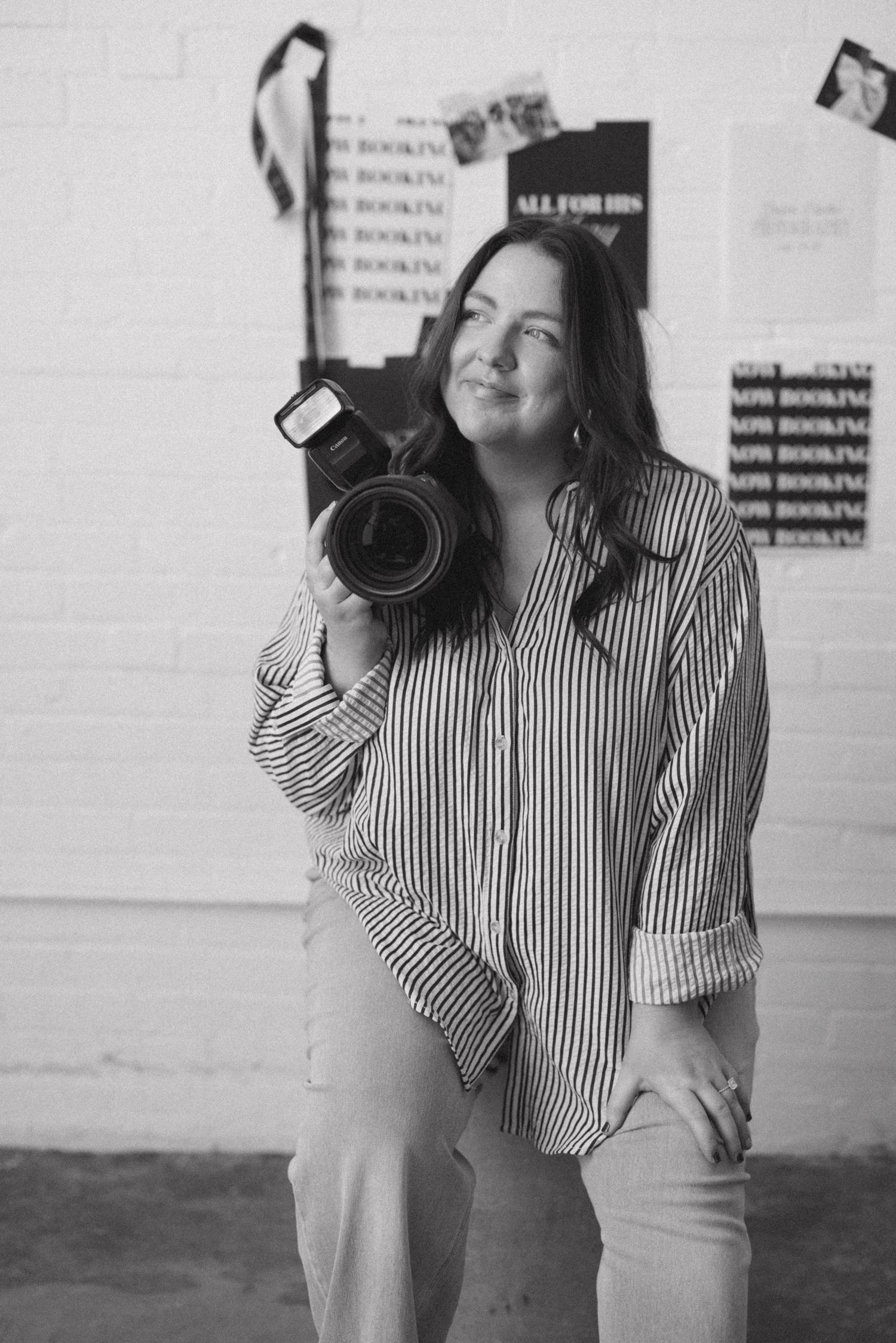 A photographer in a striped shirt takes multiple poses with a professional camera against a white wall.