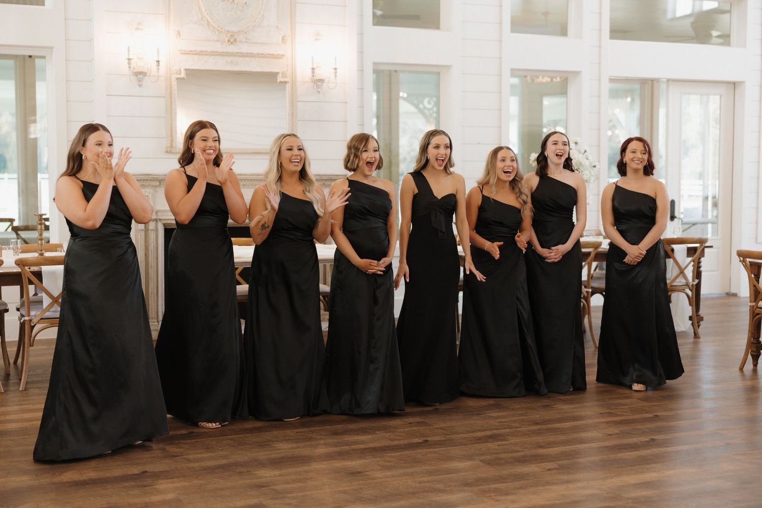 Wedding party members in black dresses pose in a bright white venue for a panoramic group photo.