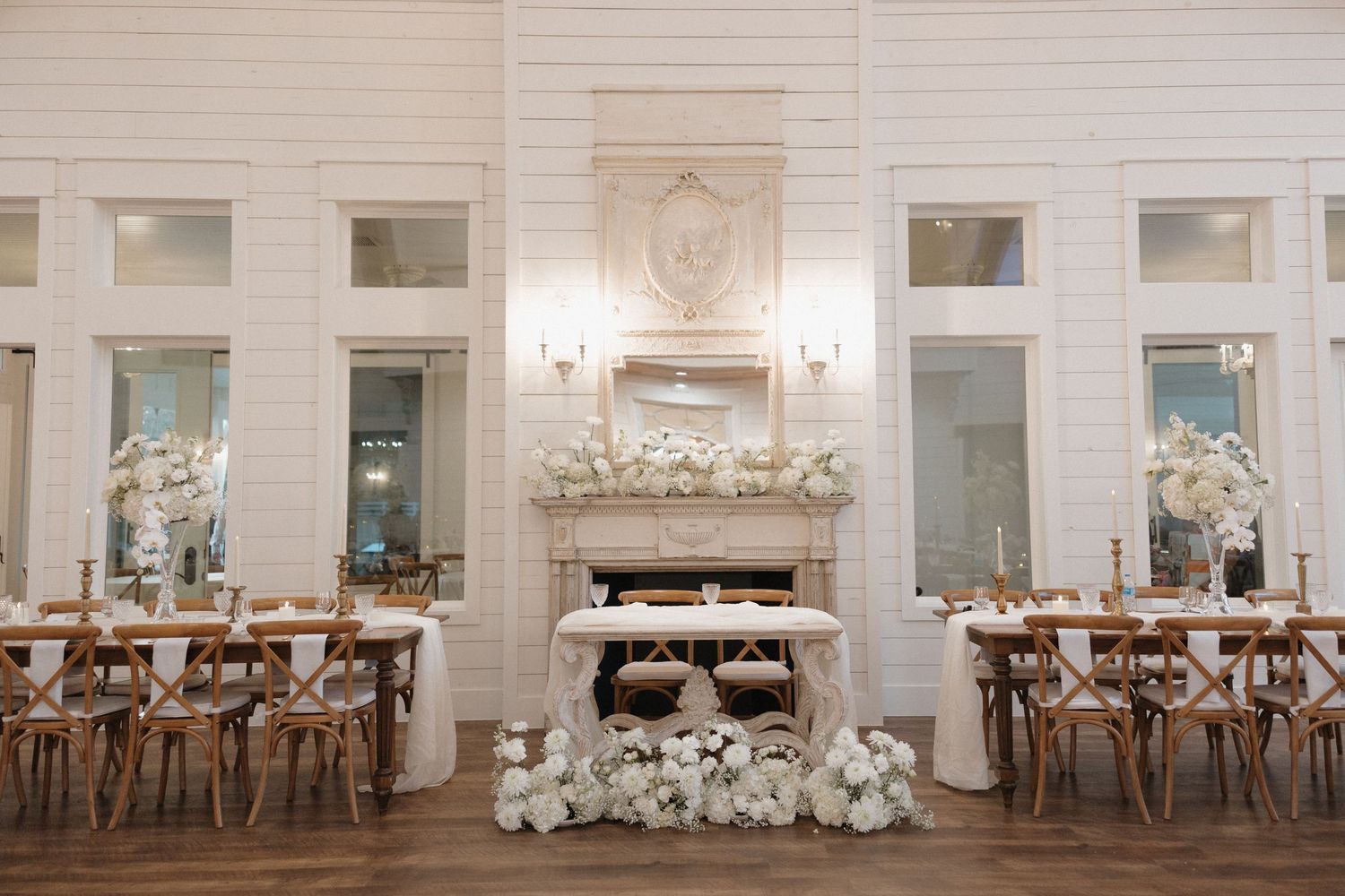 Elegant wedding reception venue with white brick walls, wooden cross-back chairs, and white floral centerpieces.