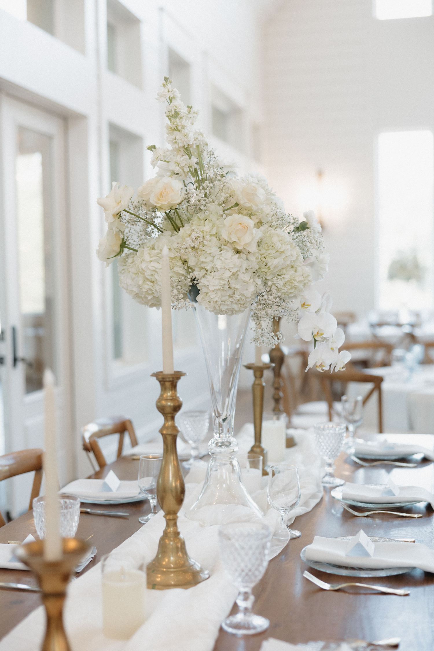 Elegant white hydrangea and floral centerpiece on gold candlestick at wedding reception table with place settings.