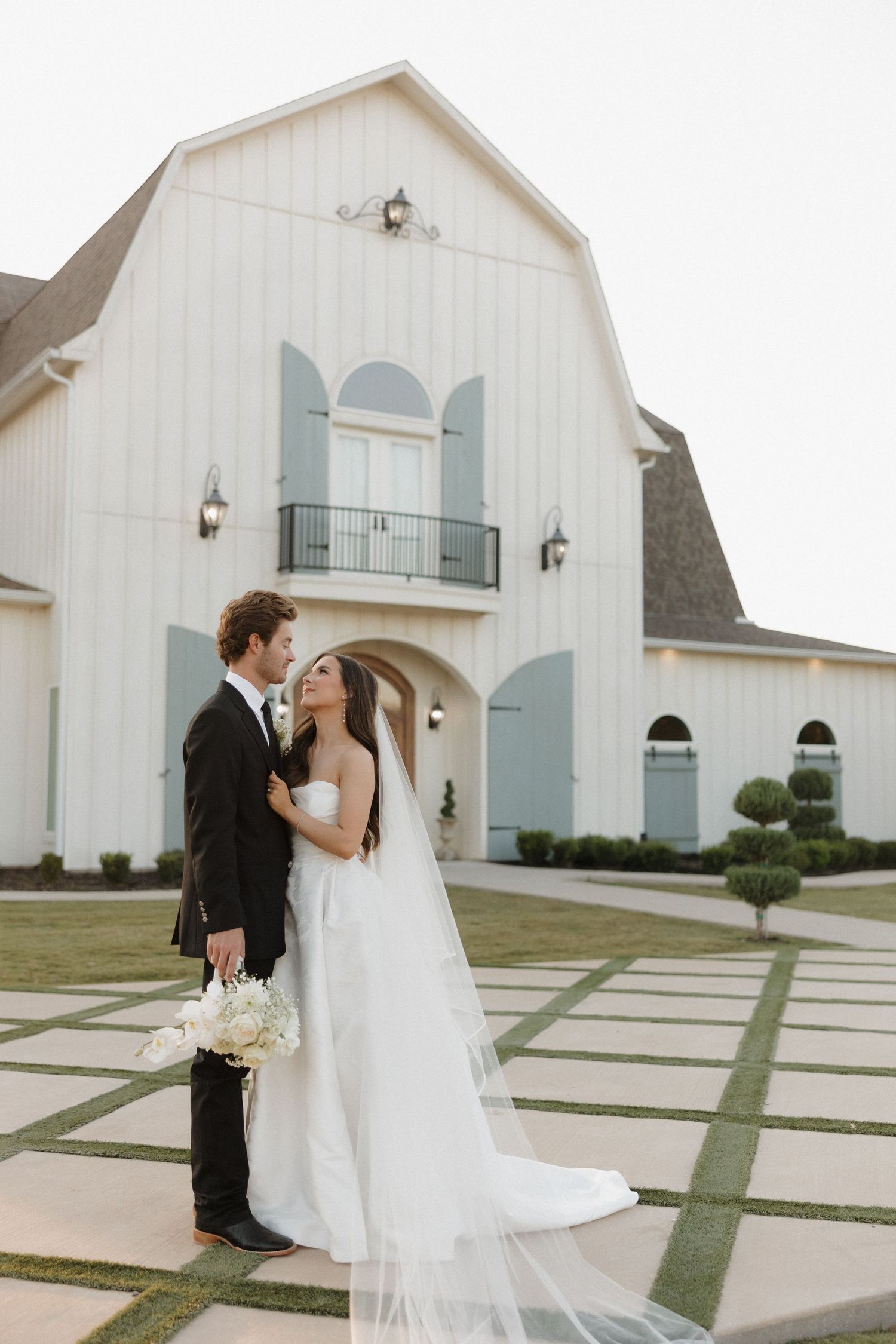 A romantic wedding couple stands outside an elegant white barn venue with pale blue shutters and a Juliet balcony.