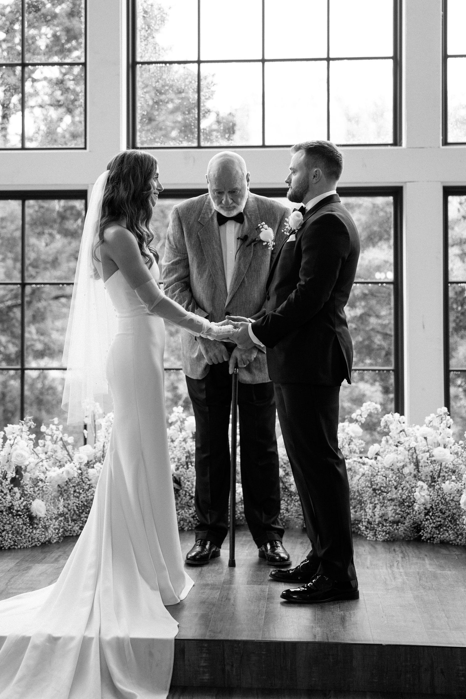 A black and white wedding ceremony takes place in front of large windows with an officiant and floral arrangements.