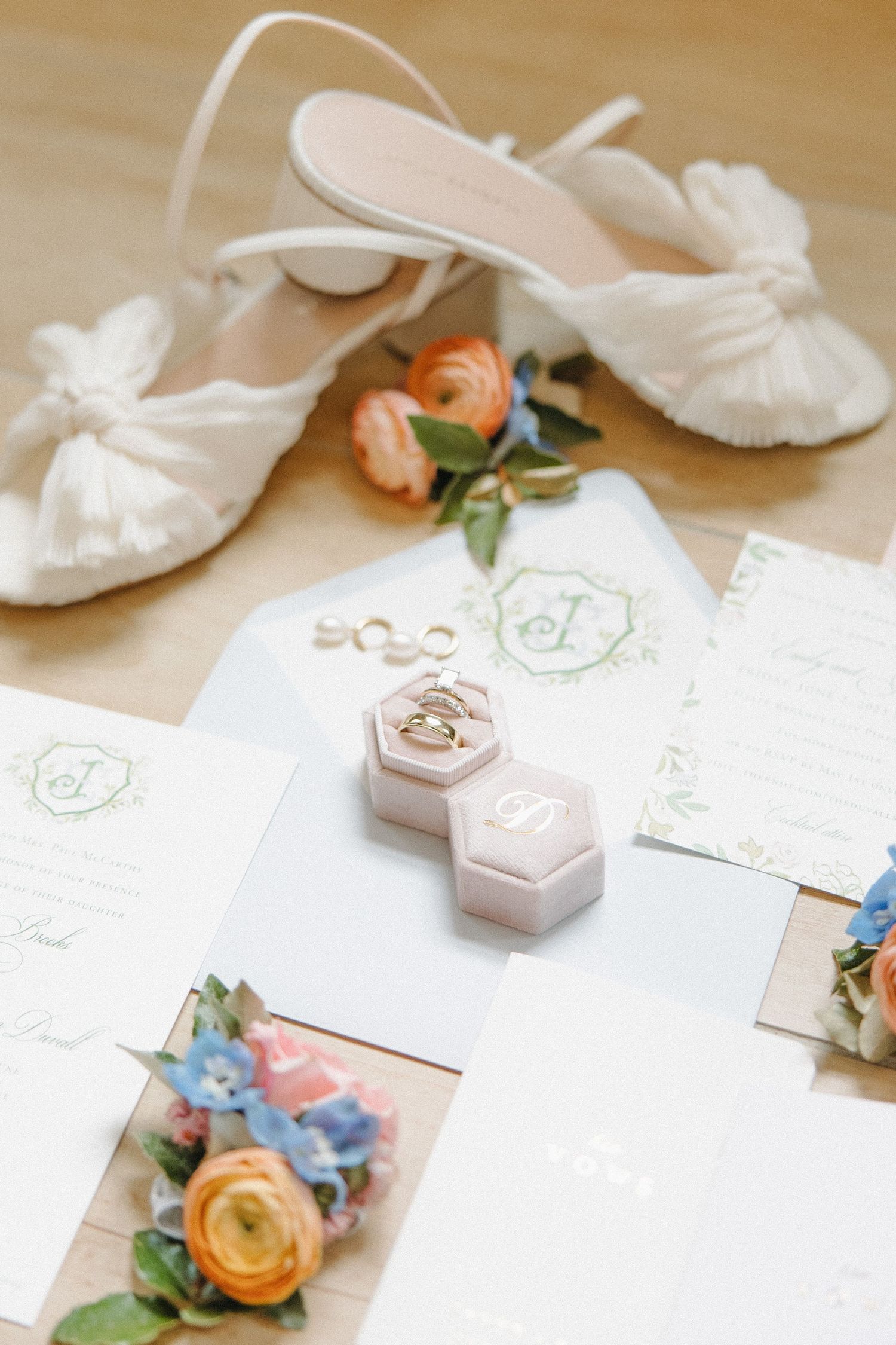 Wedding details flatlay with white bridal shoes, peach rose, invitation cards and hexagonal ring boxes on beige surface.