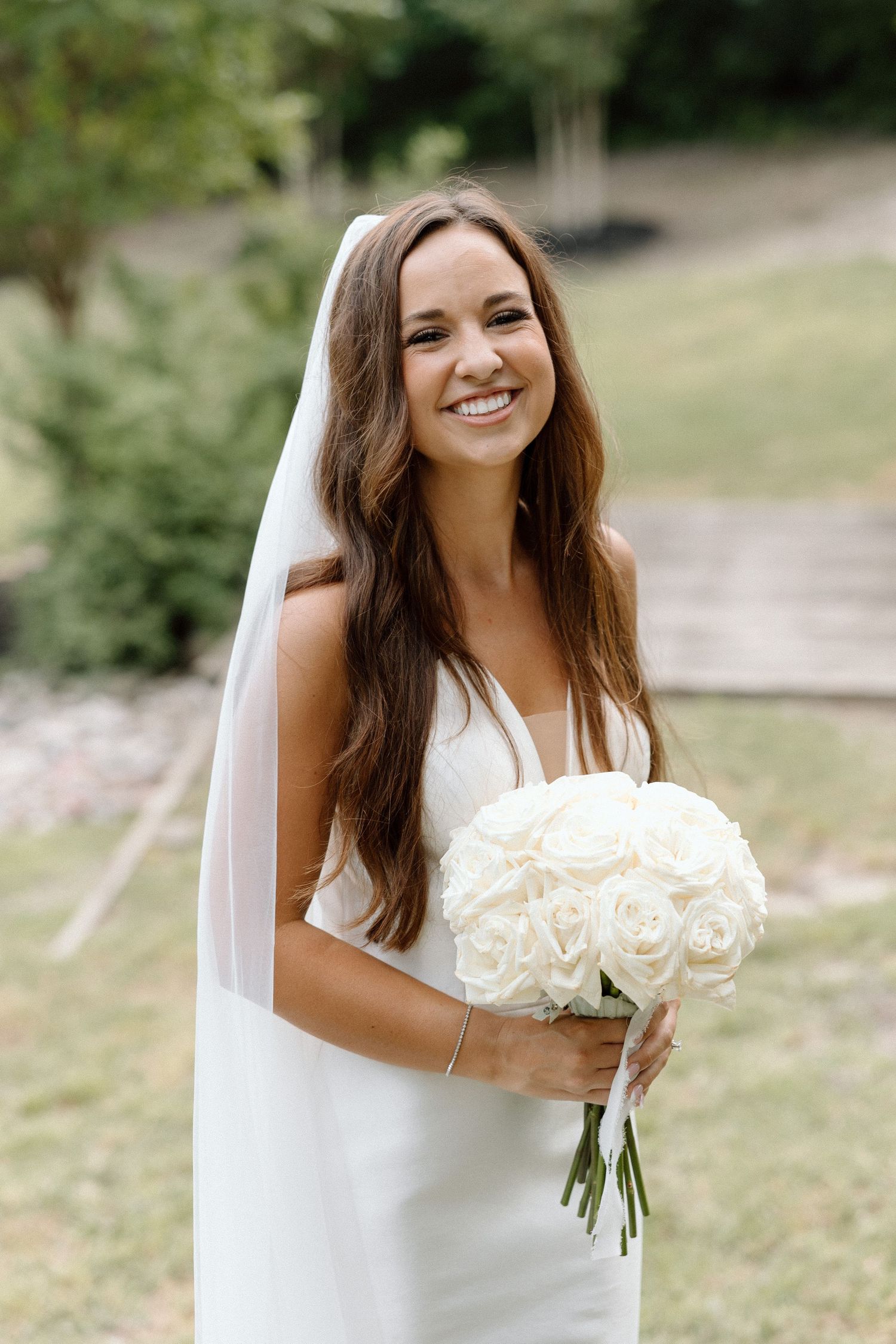 A bride in a white wedding dress holds a bouquet of white roses while posing outdoors in a natural setting.