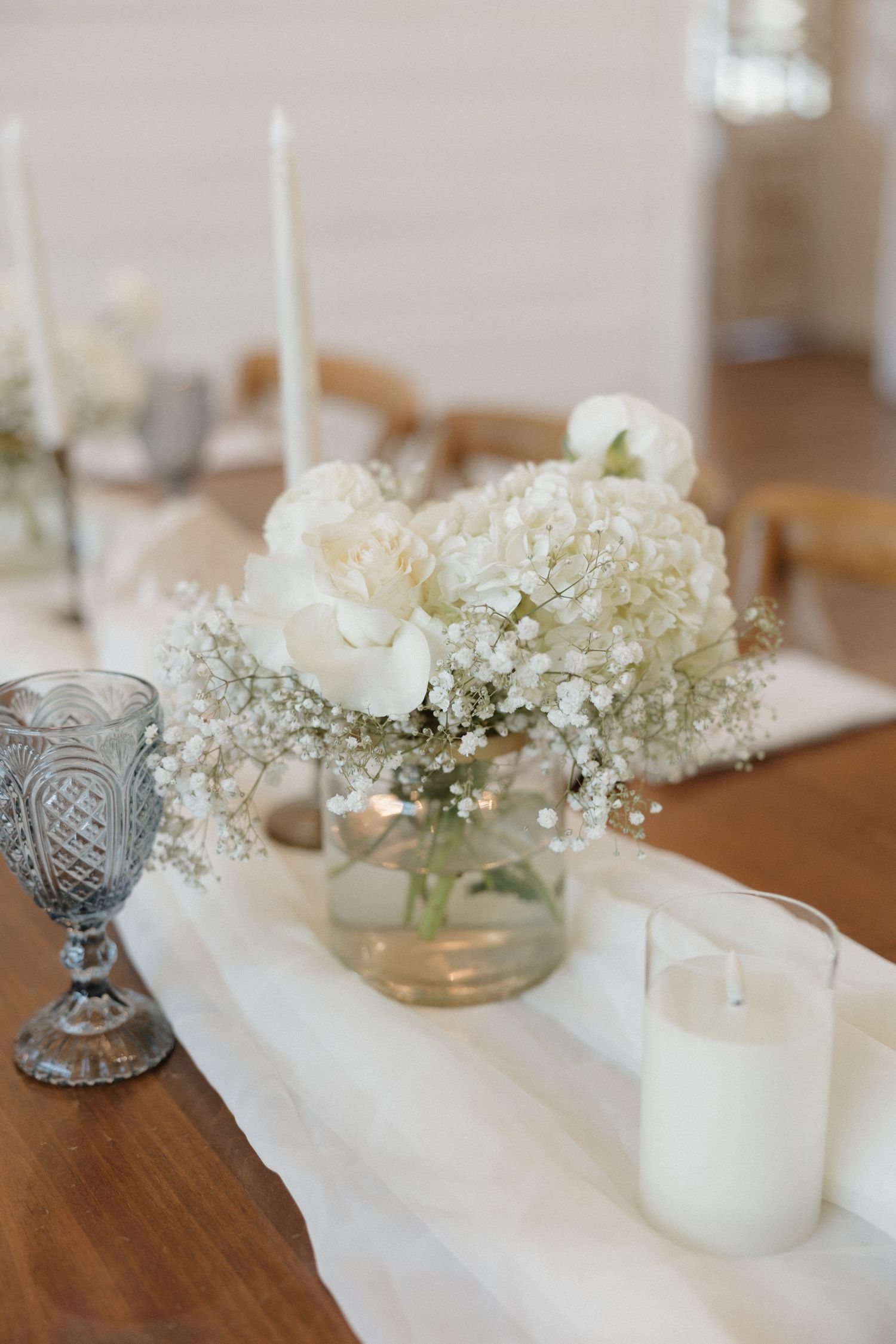 White floral centerpieces with baby's breath and roses in glass vases line a wooden table with blue vintage glassware.