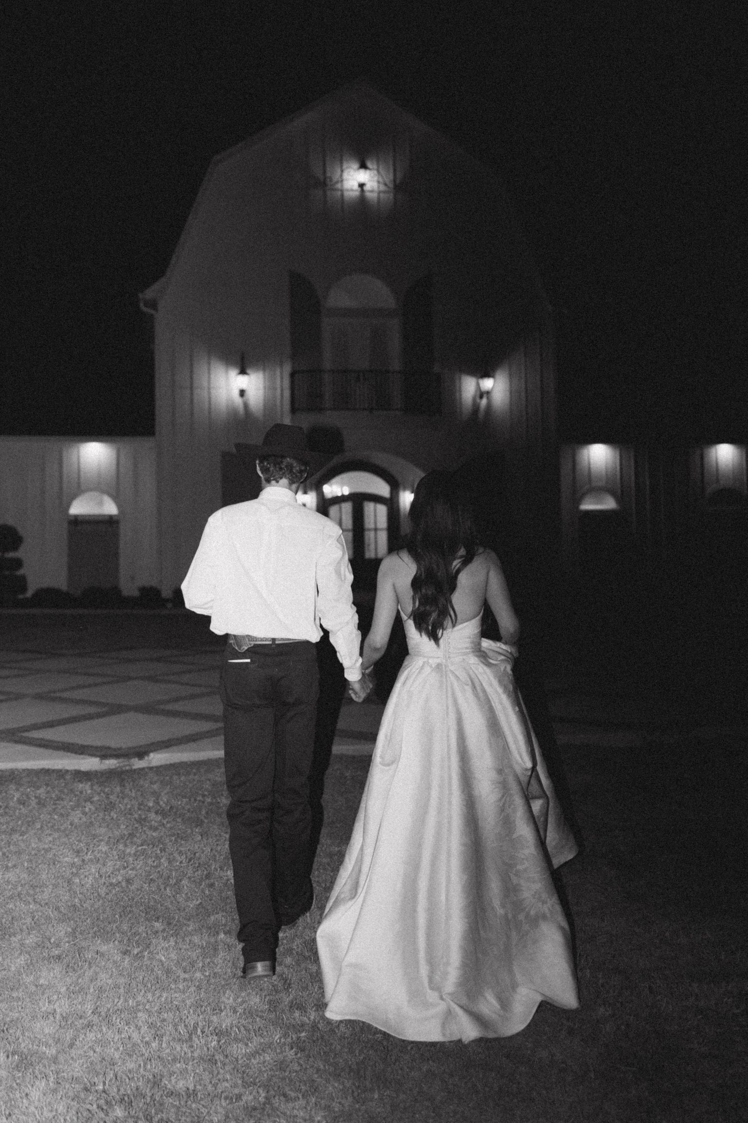 A couple in wedding attire walk hand in hand at night toward an illuminated building with glowing exterior lights.