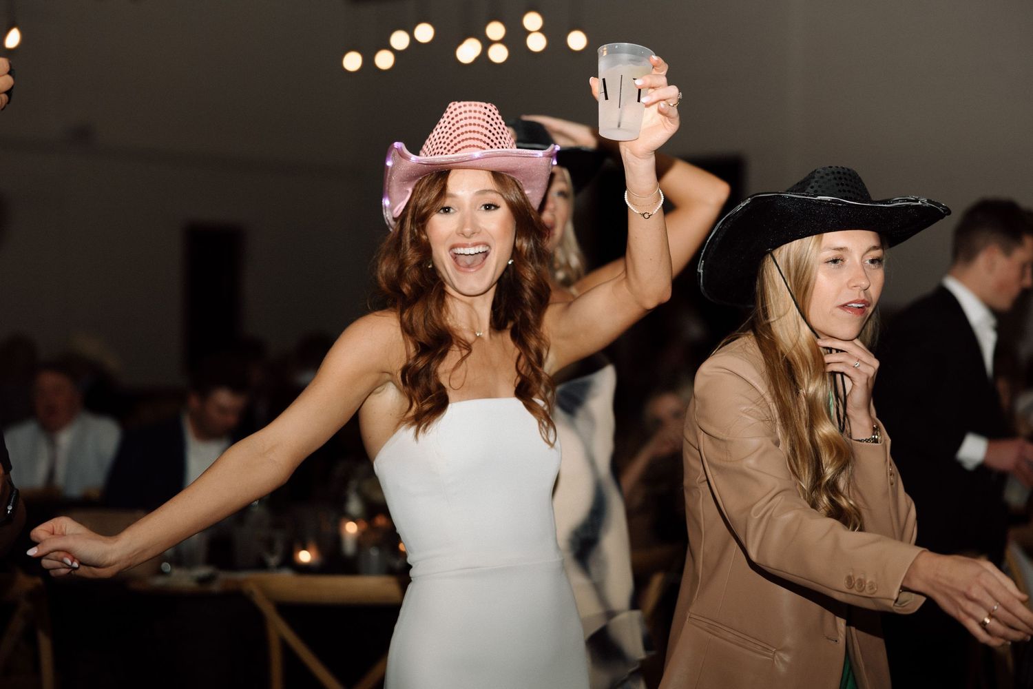 Wedding guests dance and celebrate together on a dimly lit dance floor with glowing lights in the background.
