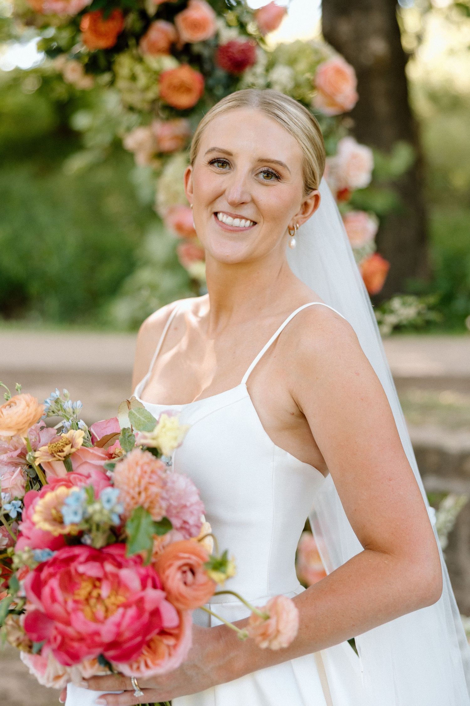 Bride in white dress holding vibrant coral and pink wedding bouquet with peach roses and greenery in outdoor garden setting.