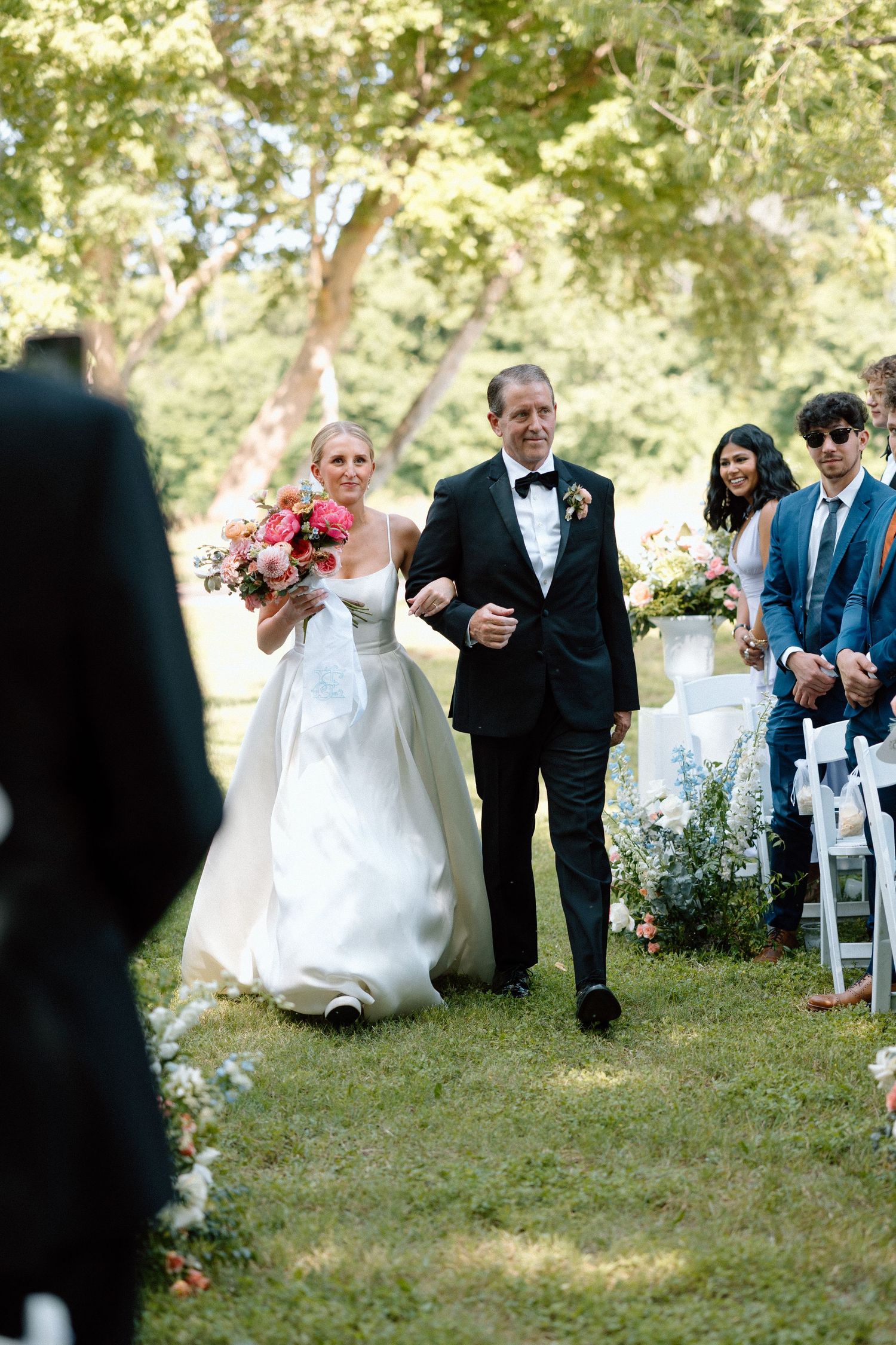 Wedding guests seated outdoors on white chairs under trees during an outdoor ceremony.