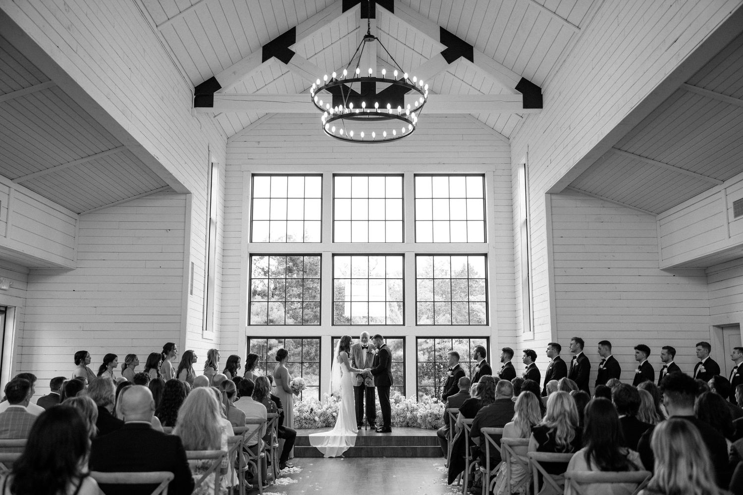 Black and white photo of a wedding ceremony inside a bright chapel with large windows and guests seated in rows.
