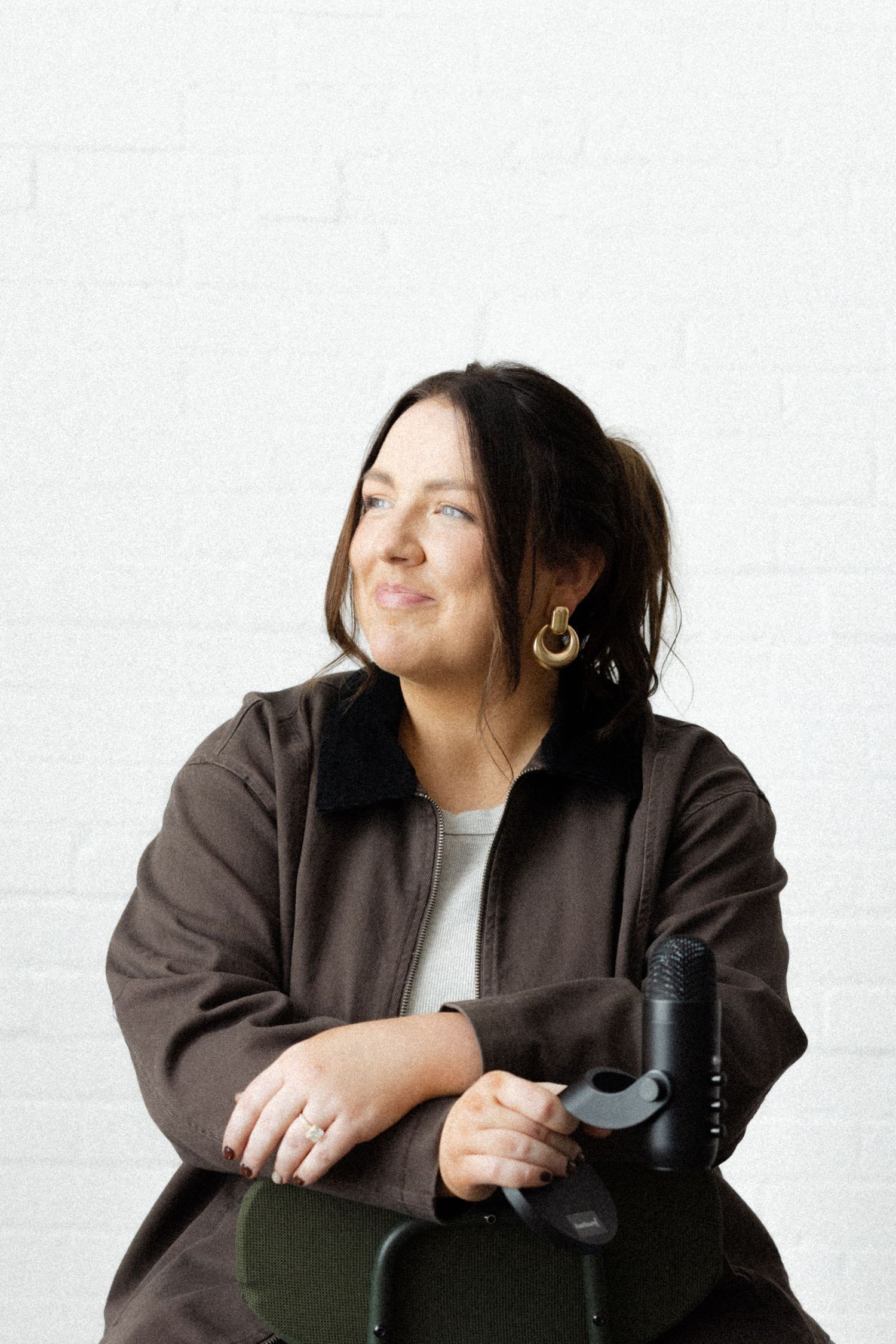 Woman in brown jacket with gold earrings sits relaxed against white brick wall, smiling thoughtfully with microphone nearby.