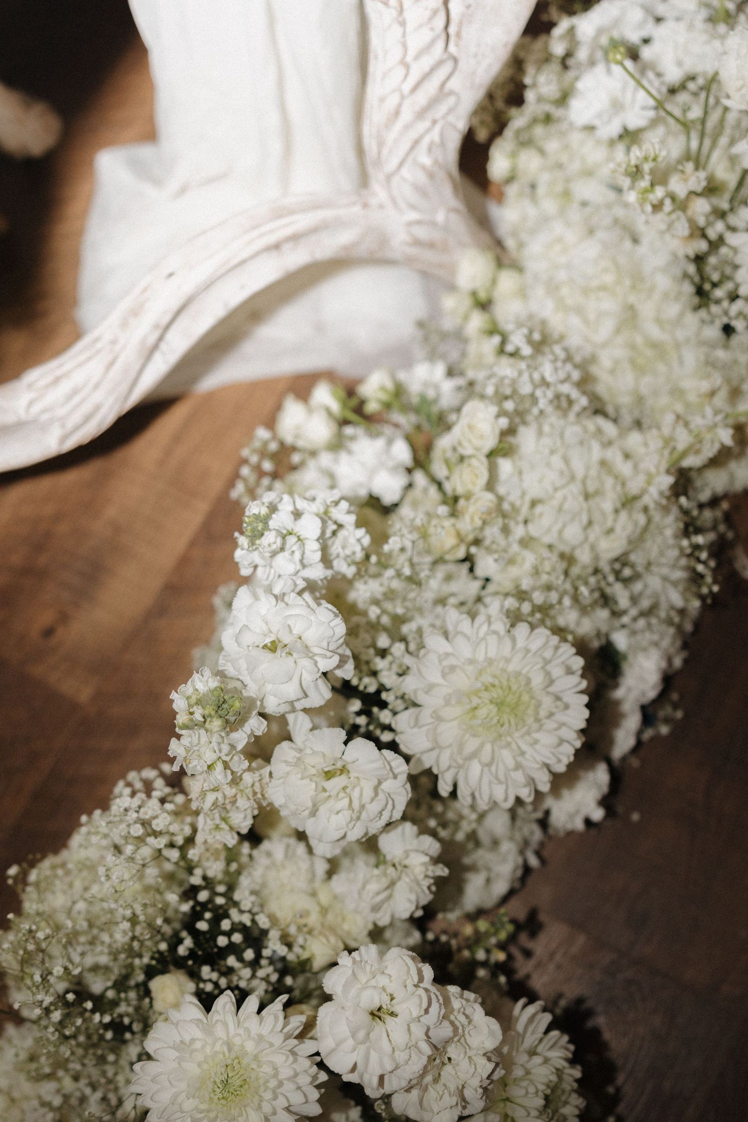 White chrysanthemums and baby's breath flowers arranged on wooden surface with bridal shoe in background.