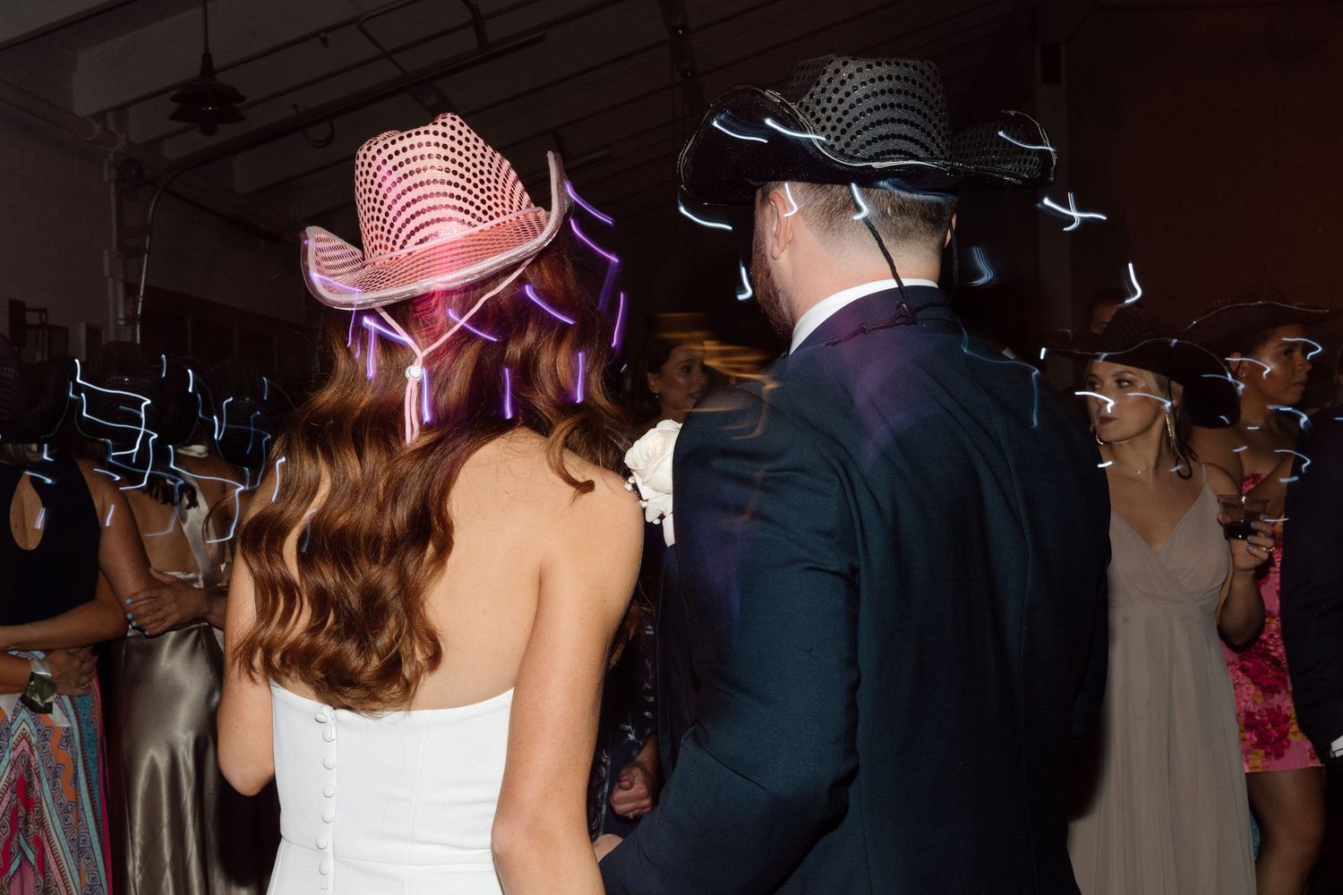 Wedding guests dance together on a dark dance floor illuminated by blue lights at an evening reception.