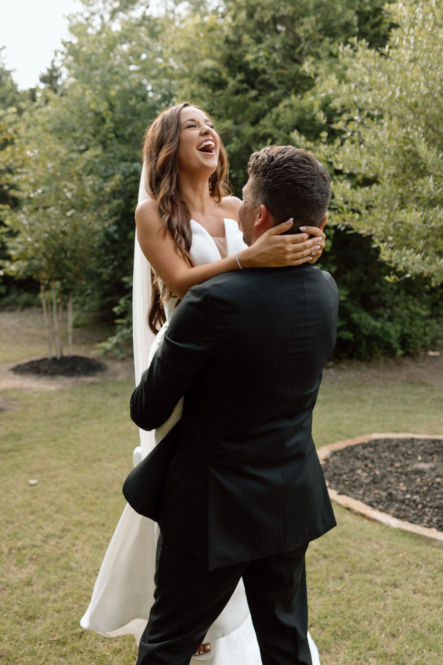 A couple in wedding attire embraces joyfully outdoors in a garden setting with trees in the background.