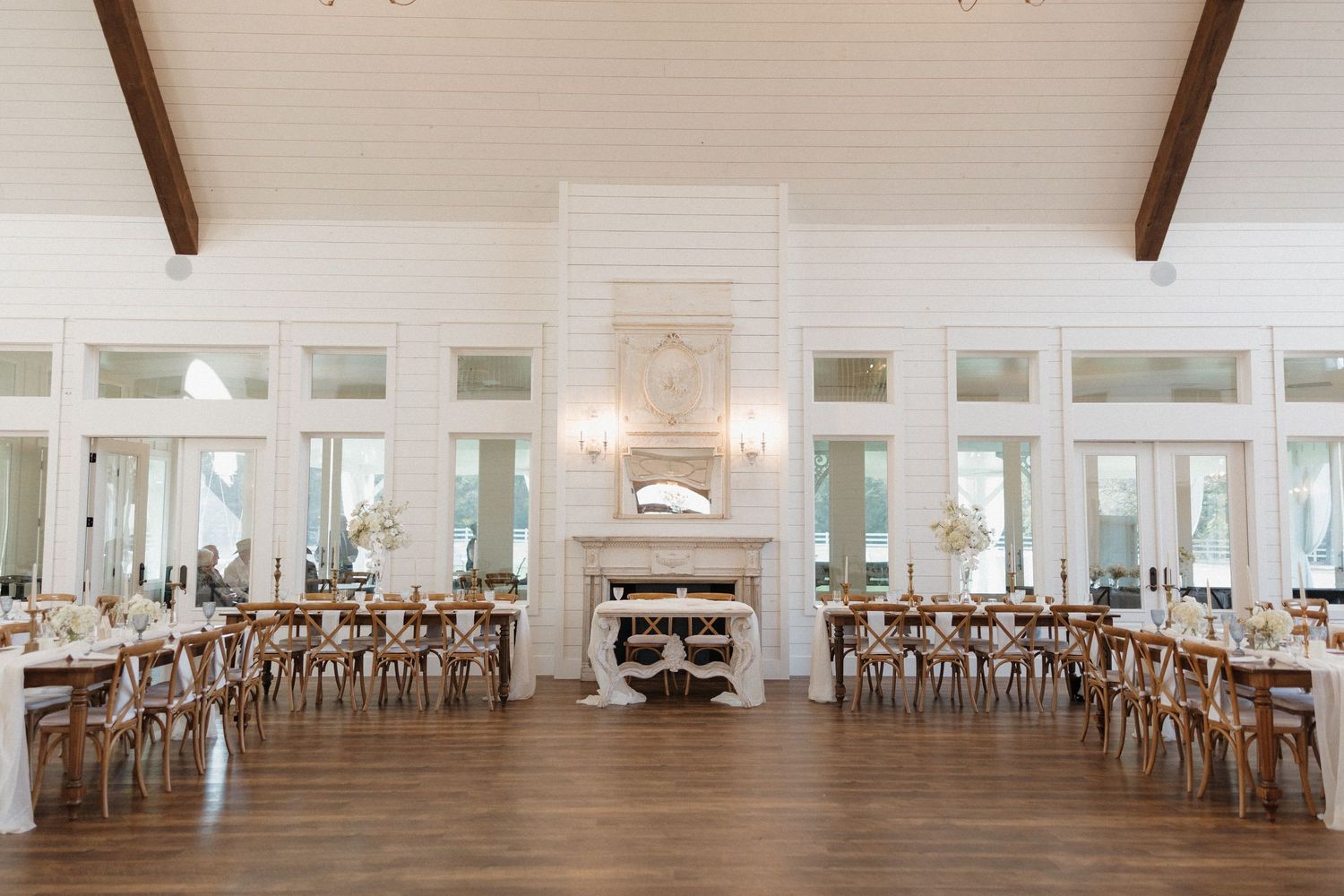A bright white wedding reception venue with elegant chandeliers and wooden cross-back chairs arranged in rows.