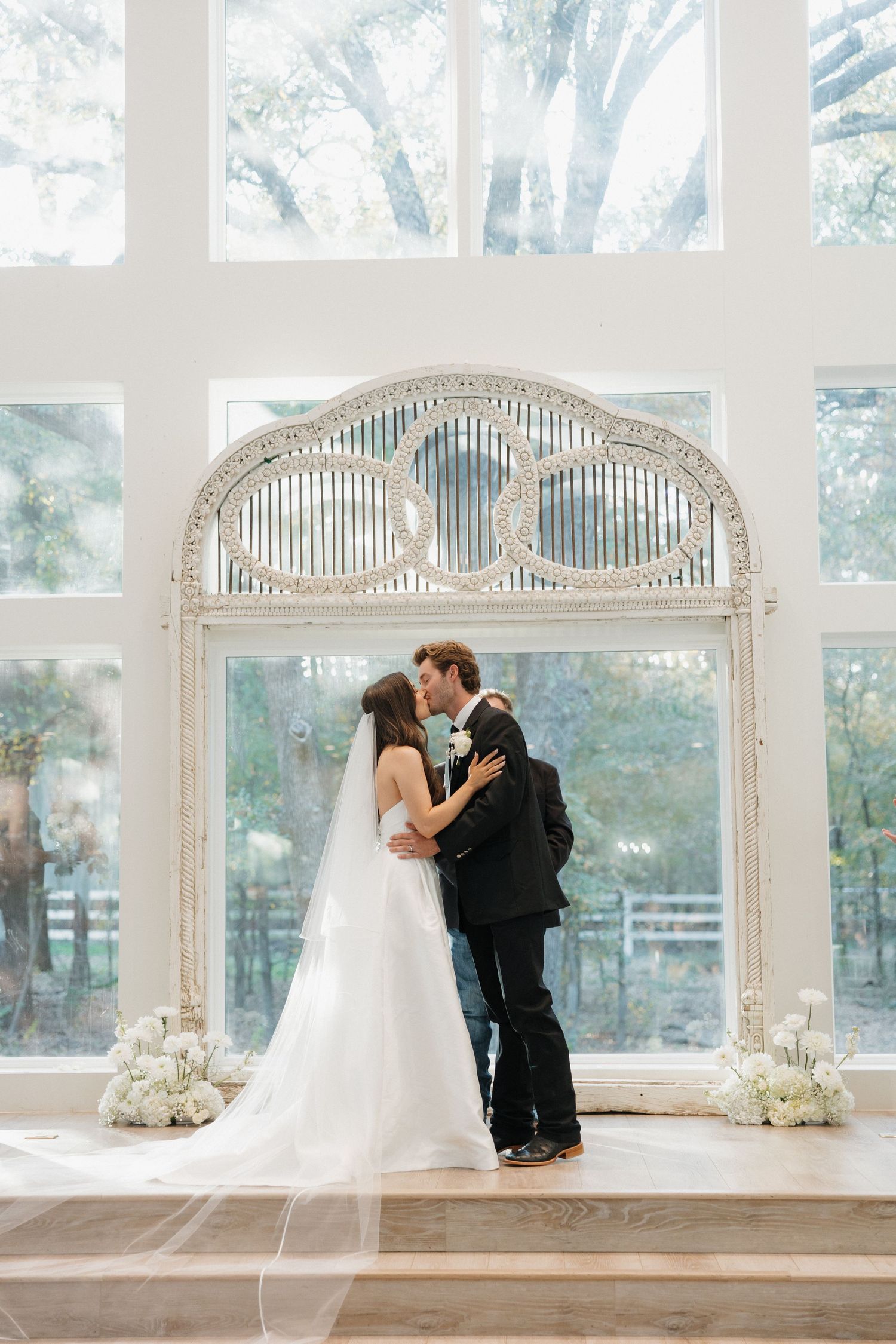 A romantic wedding scene showing a couple sharing a kiss under elegant white arched windows in a bright sunlit room.