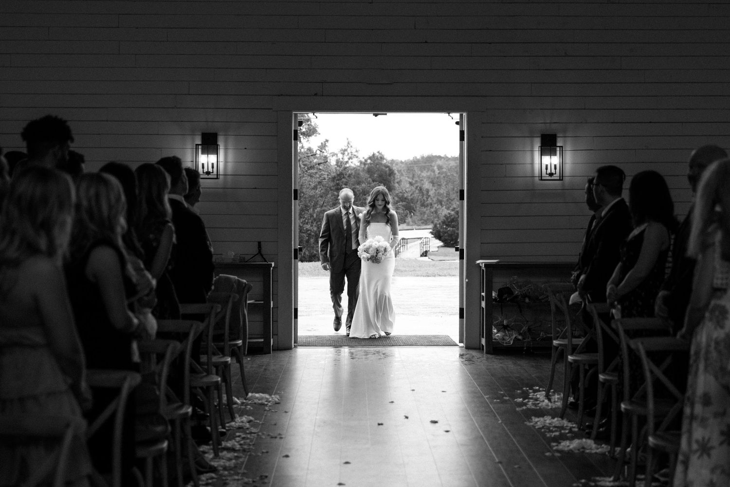A silhouetted couple stands in a barn doorway at a wedding ceremony while guests line the aisle in black and white.