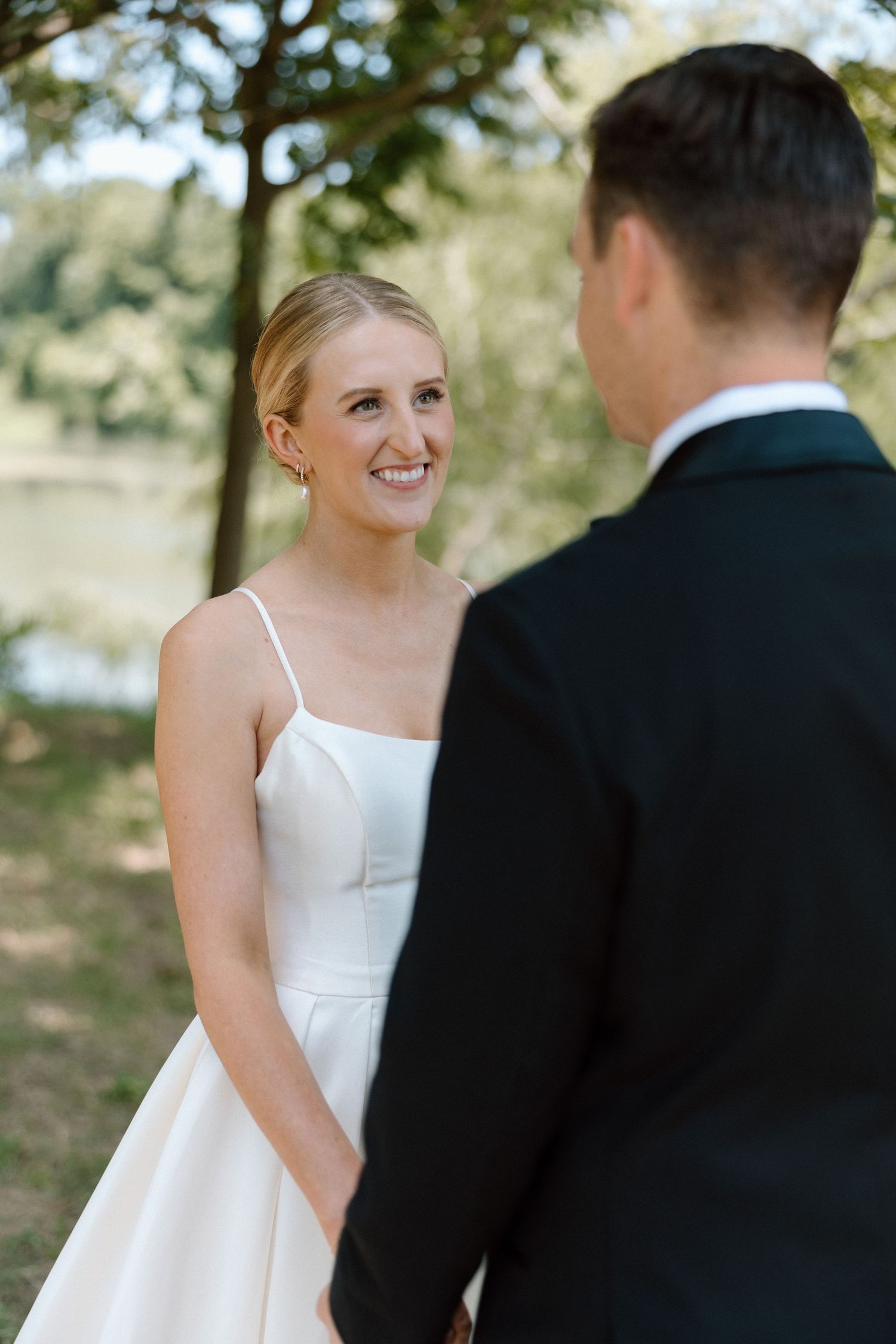 A bride in a white spaghetti strap wedding dress shares a joyful moment with someone wearing a black suit outdoors.