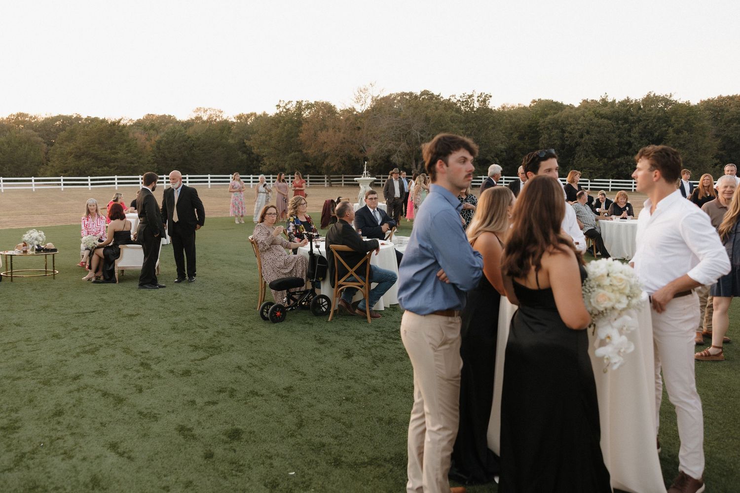Guests mingle on a grassy field at an outdoor event during sunset with white fencing in the background.