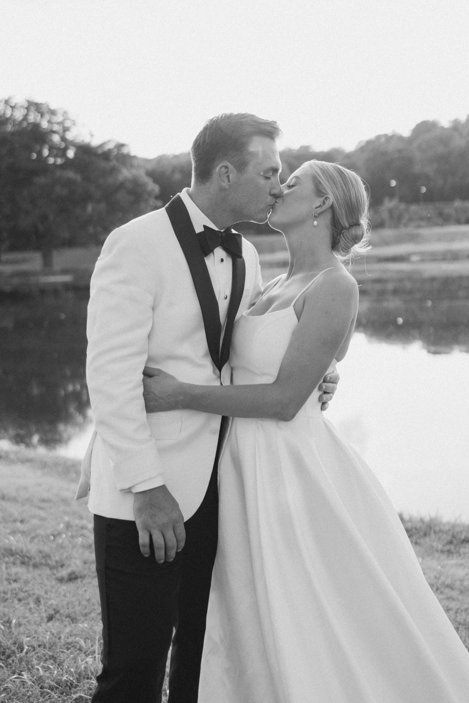 A couple shares a romantic kiss outdoors by a lake in formal wedding attire.