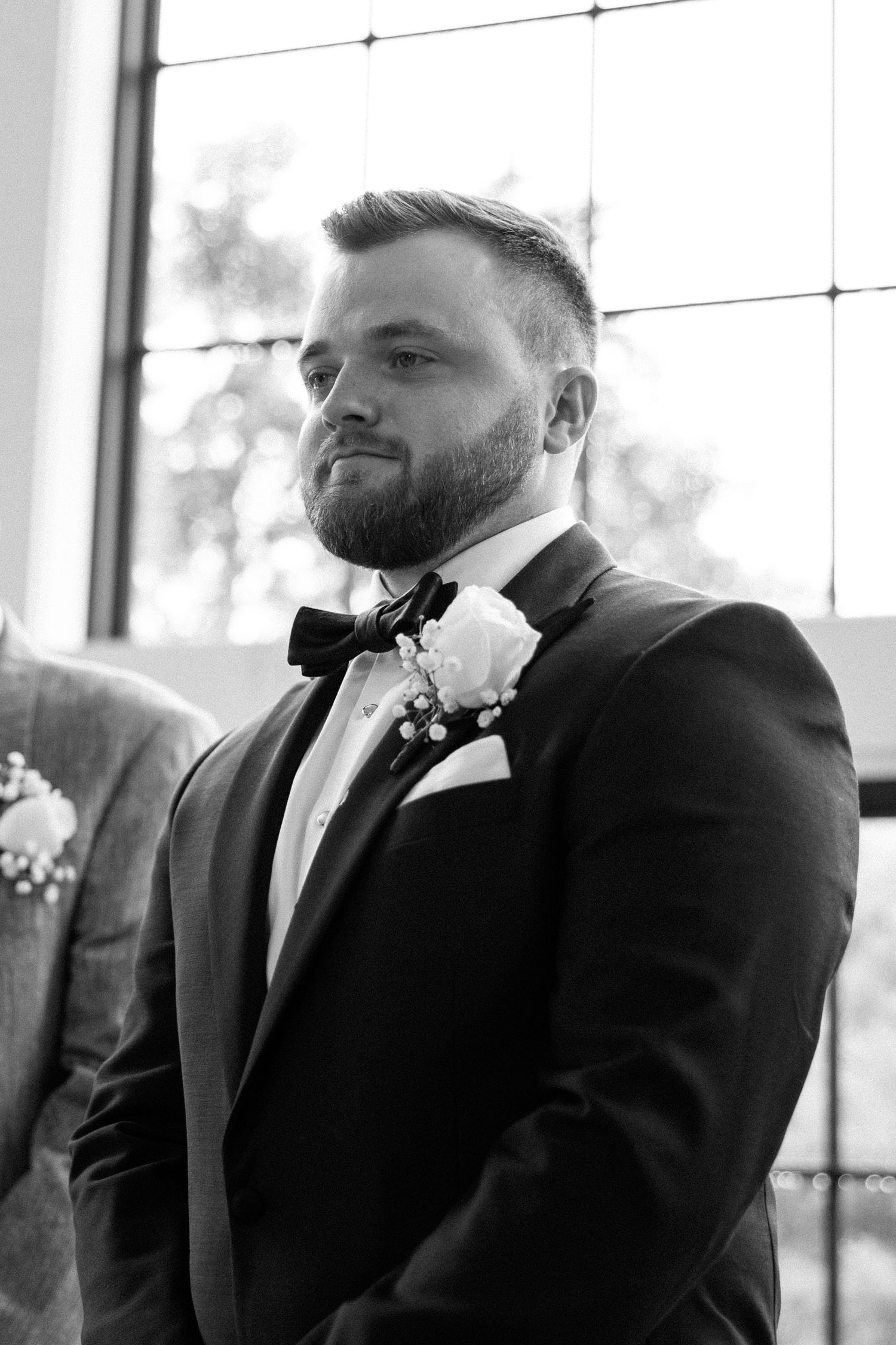 Black and white wedding photo showing a groom in a classic tuxedo with bow tie and boutonniere.
