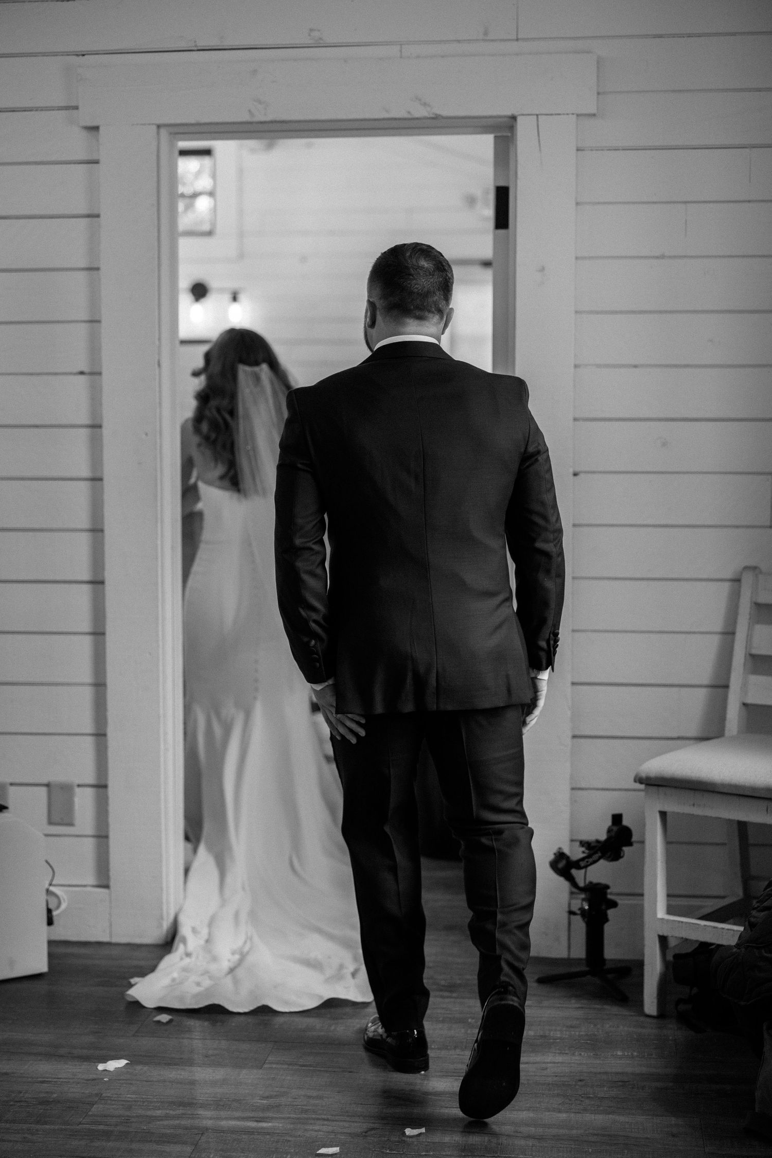 Black and white photo of a bride and groom walking through a doorway in a rustic white-walled interior.