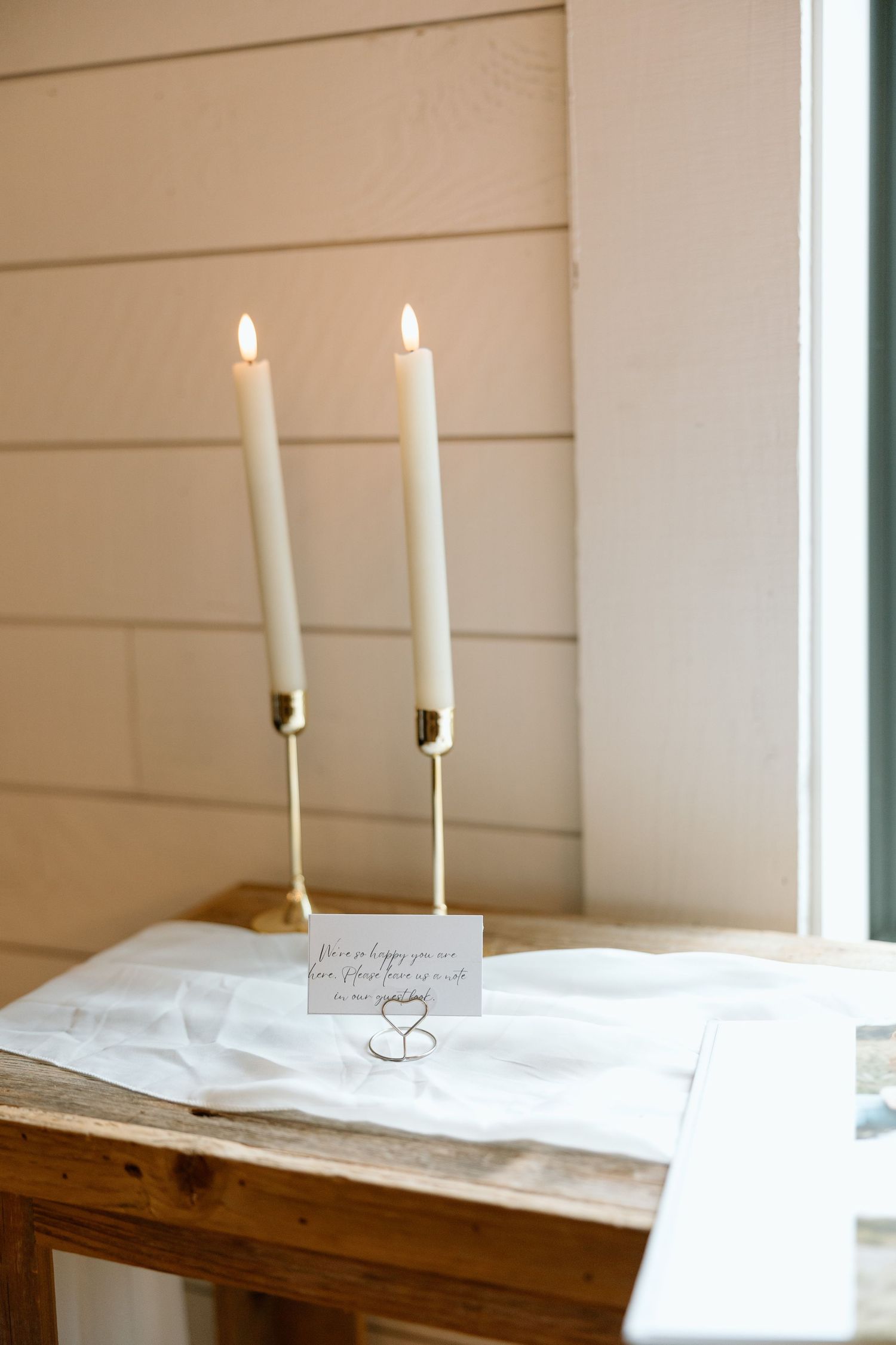 Two white candles burning on a rustic wooden table with a white tablecloth.