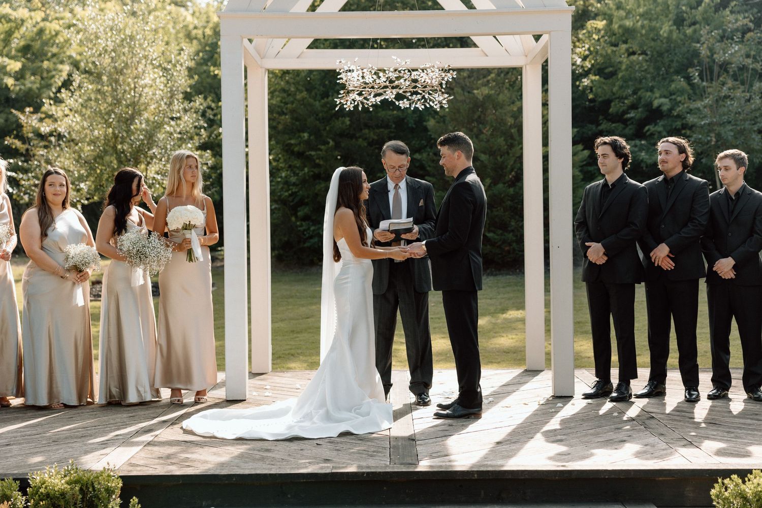 Wedding party stands in elegant formation under white gazebo during outdoor ceremony.
