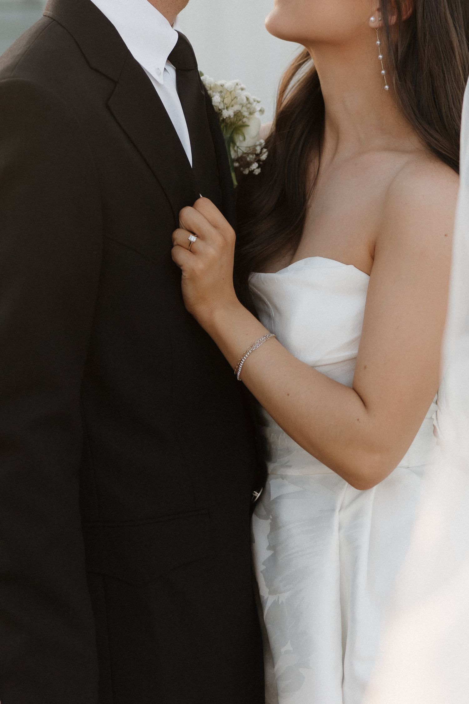 Close-up of bride in white strapless gown and groom in black suit with boutonniere embracing during wedding photoshoot.