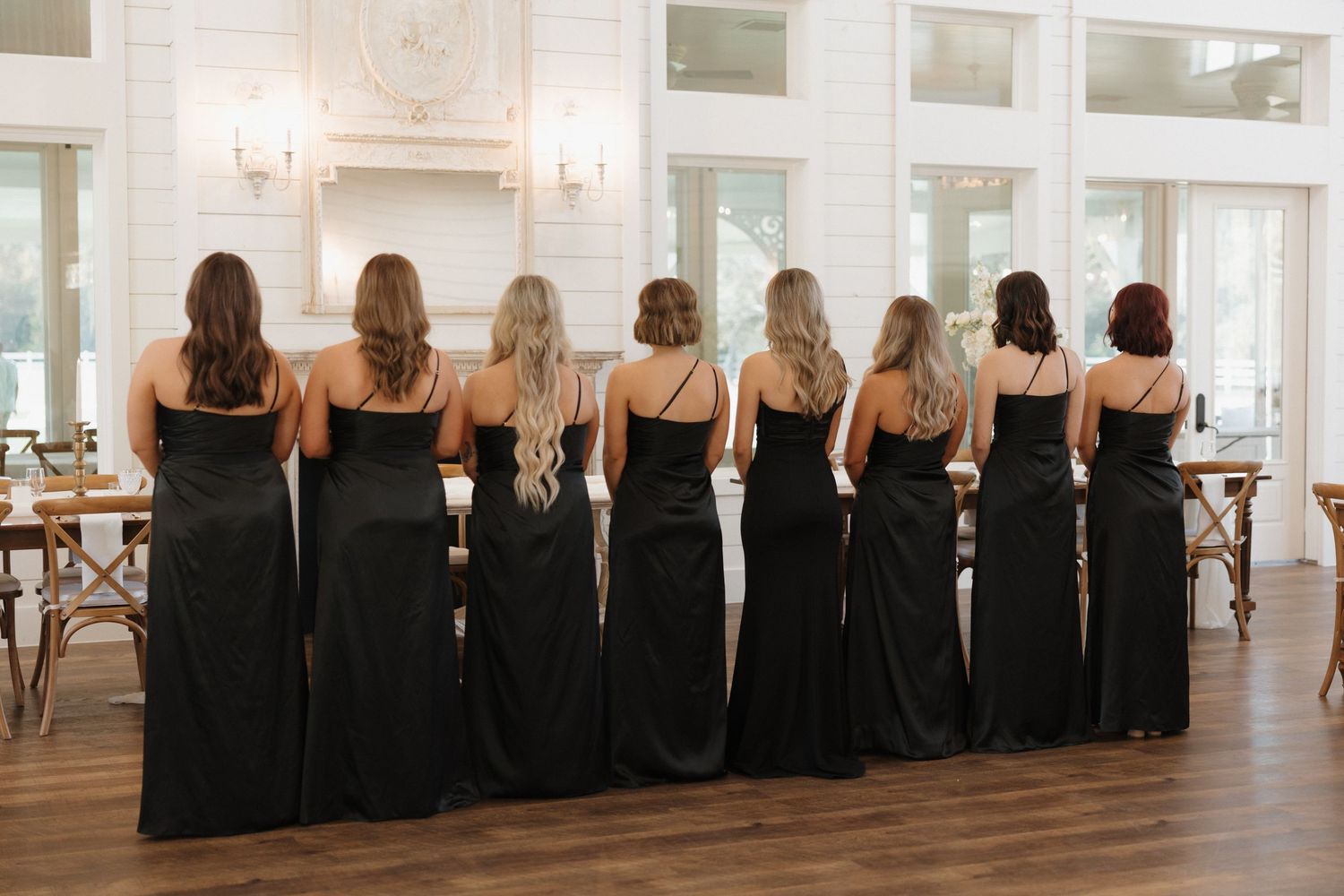 A row of bridesmaids in matching black dresses stand together in an elegant white room.