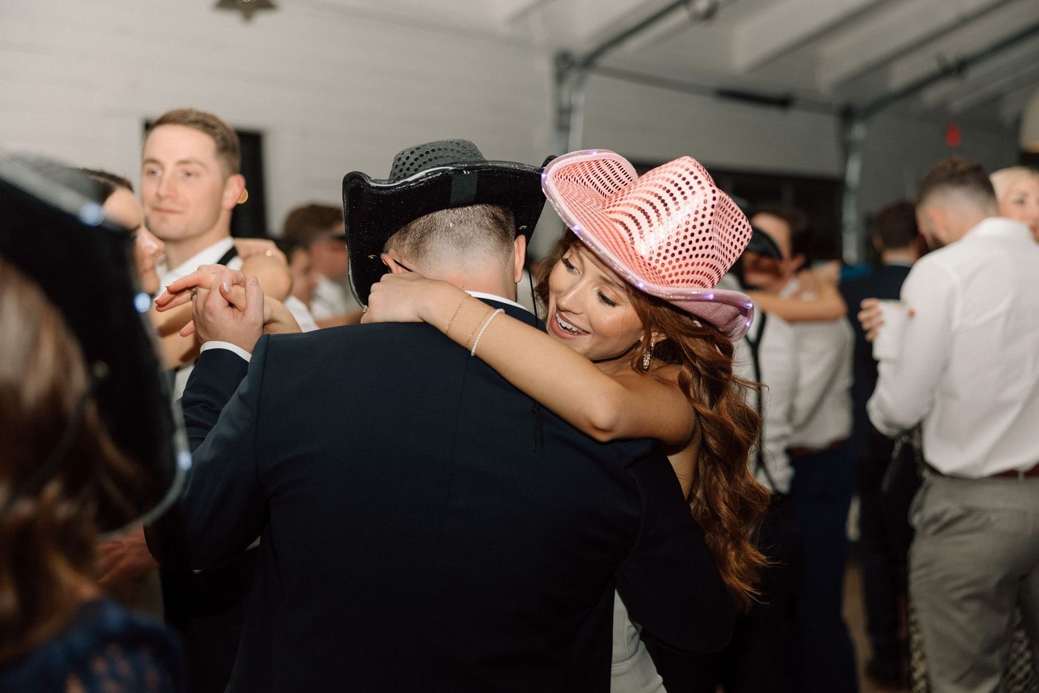 Couple dancing together at a crowded indoor reception, with the person wearing a pink hat embracing their partner.