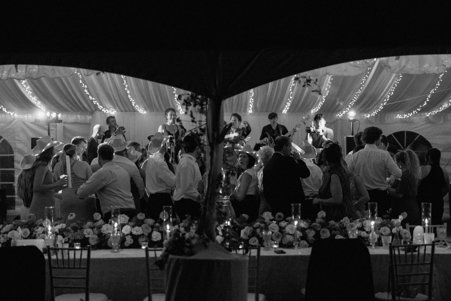 Black and white image showing wedding guests dancing under a tent decorated with string lights at night.