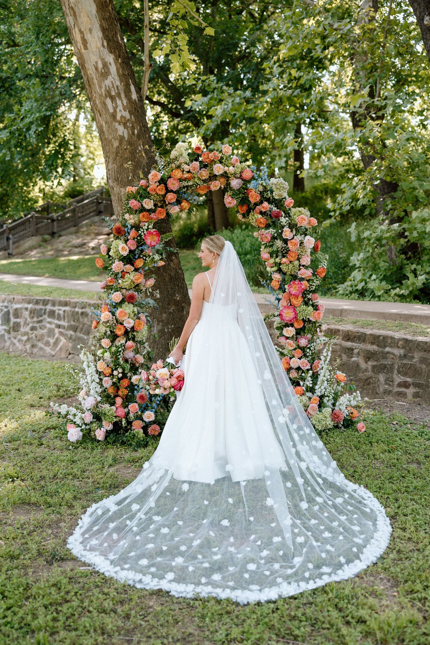 A floral wedding arch with coral and peach roses frames a long lace veil and white gown in an outdoor garden setting.