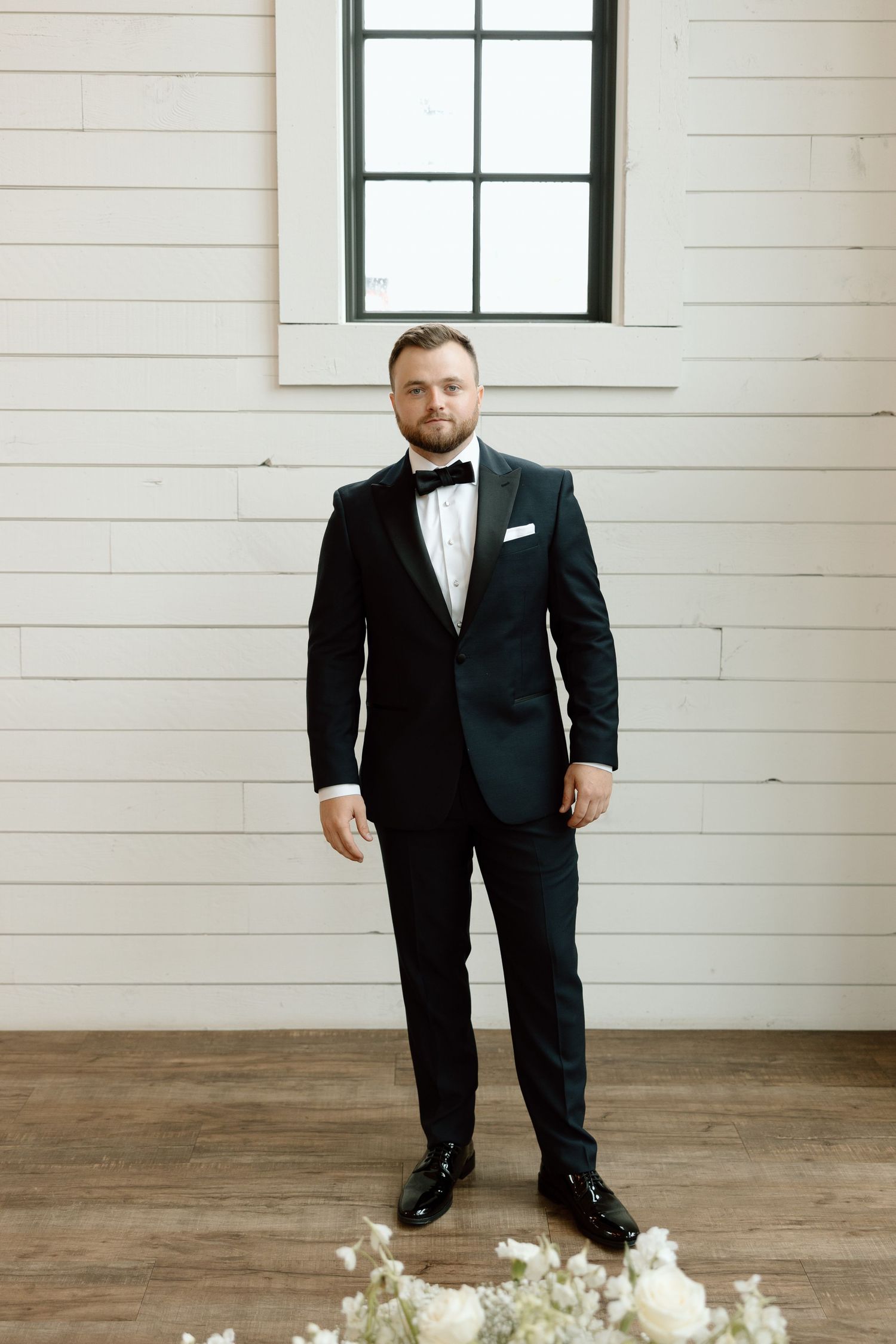 A person in a formal black tuxedo and bow tie stands in a bright white room with hardwood floors.