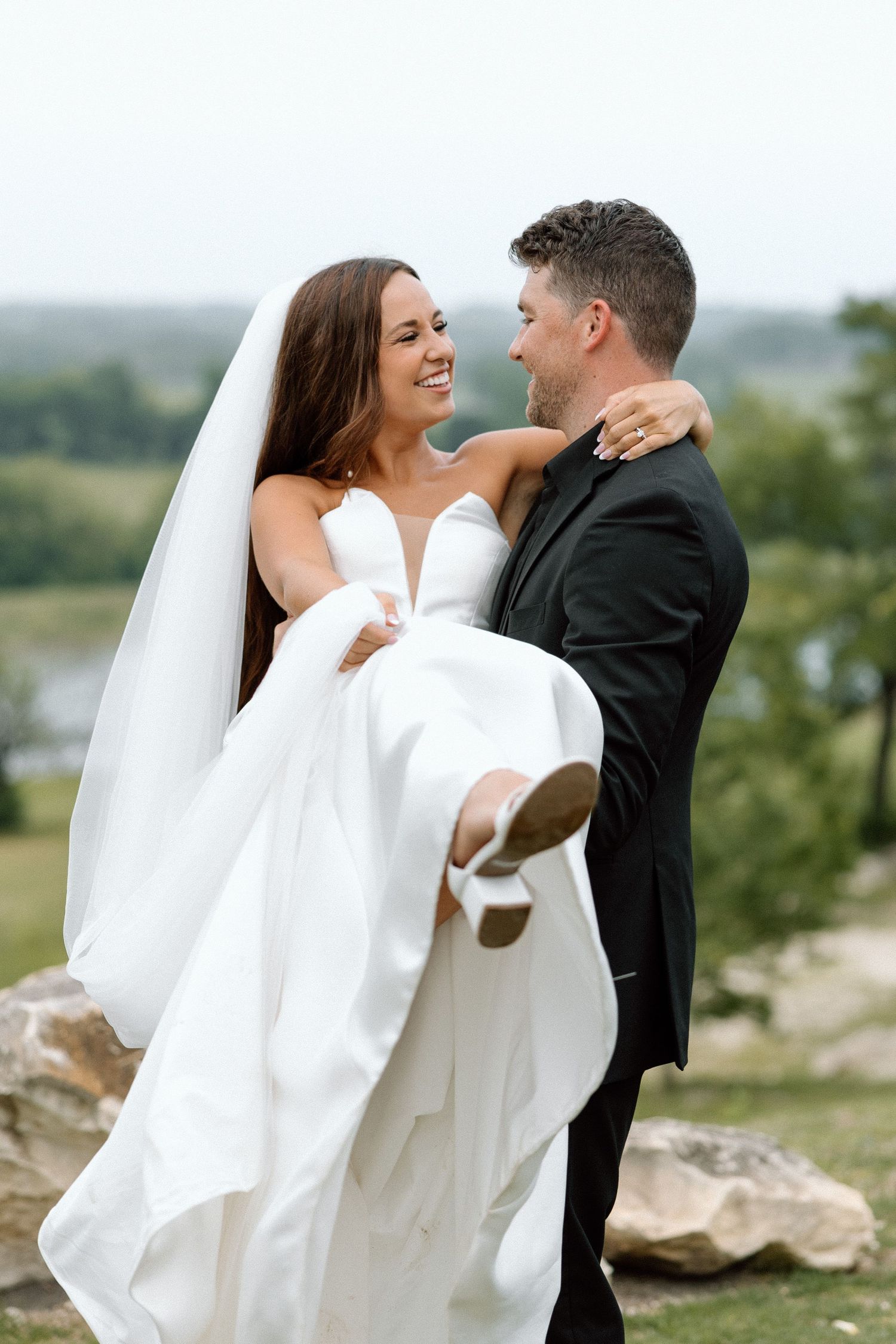 A romantic wedding couple embraces outdoors as the groom lifts the bride in her flowing white dress and veil.
