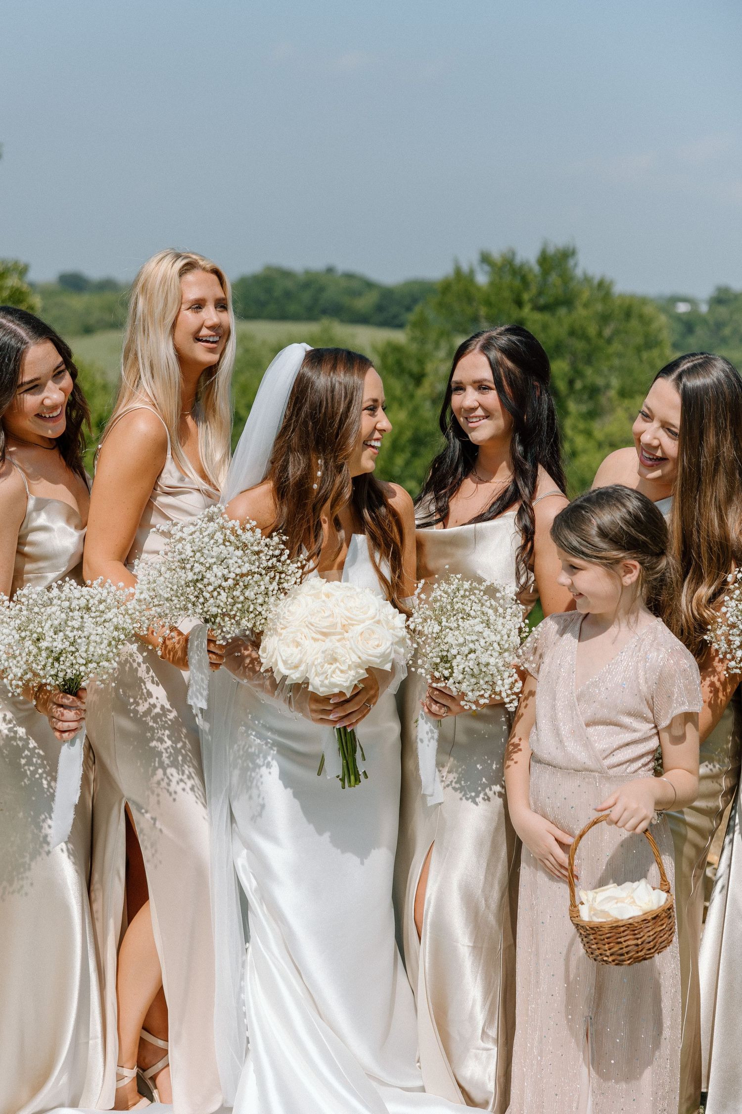 Bridesmaids in white dresses holding baby's breath bouquets laugh together outdoors on sunny day.