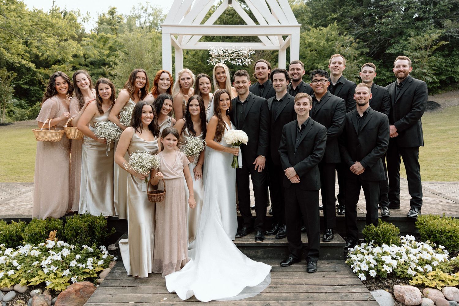 Wedding party poses together in formal attire for group photos in outdoor garden setting with white floral arrangements.
