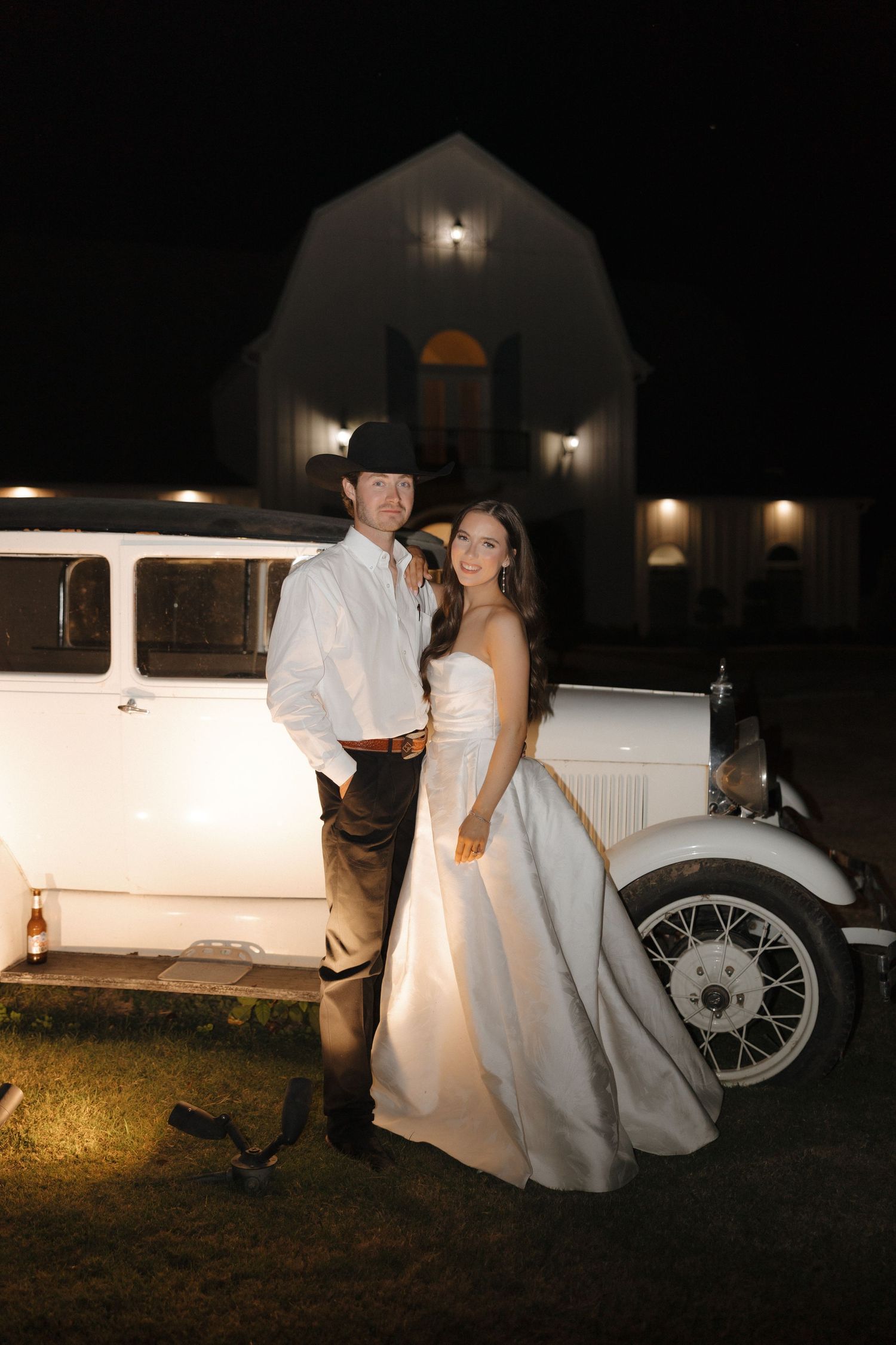 Vintage wedding scene at night with a classic white car and couple in formal attire posing by moonlight.