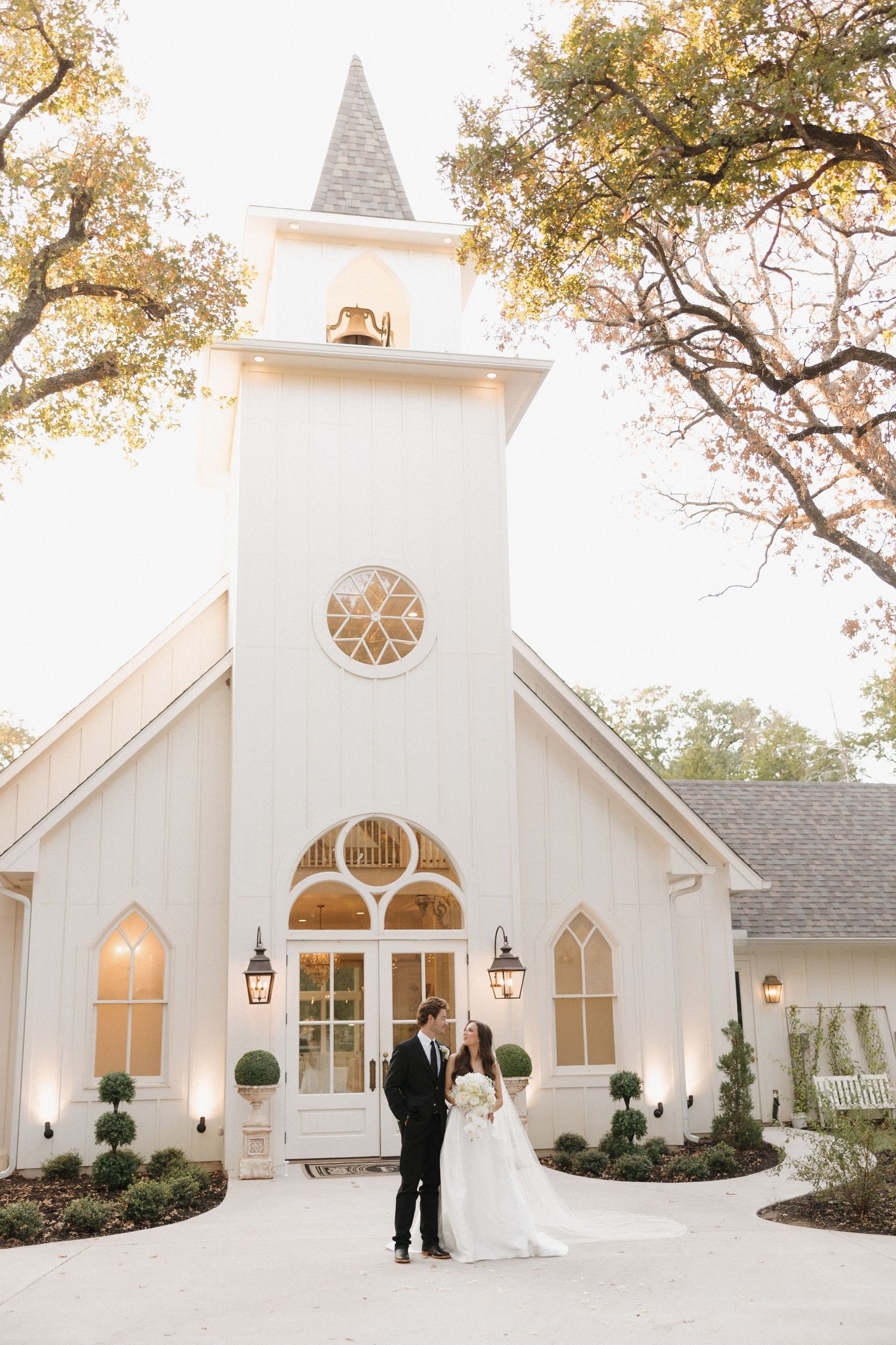 Elegant white chapel wedding venue with arched windows and steeple surrounded by trees in golden sunlight.