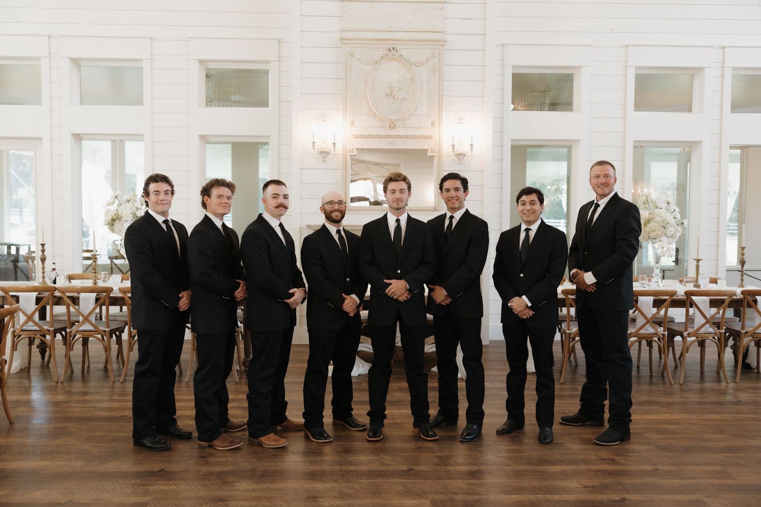 Groomsmen in black suits pose together in an elegant white church venue with large windows and wooden floors.