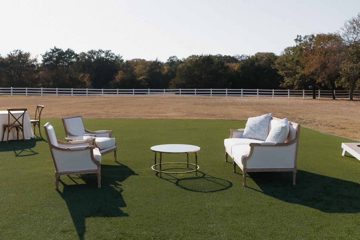Elegant outdoor lounge furniture arranged on green turf with white fencing and trees in the background.