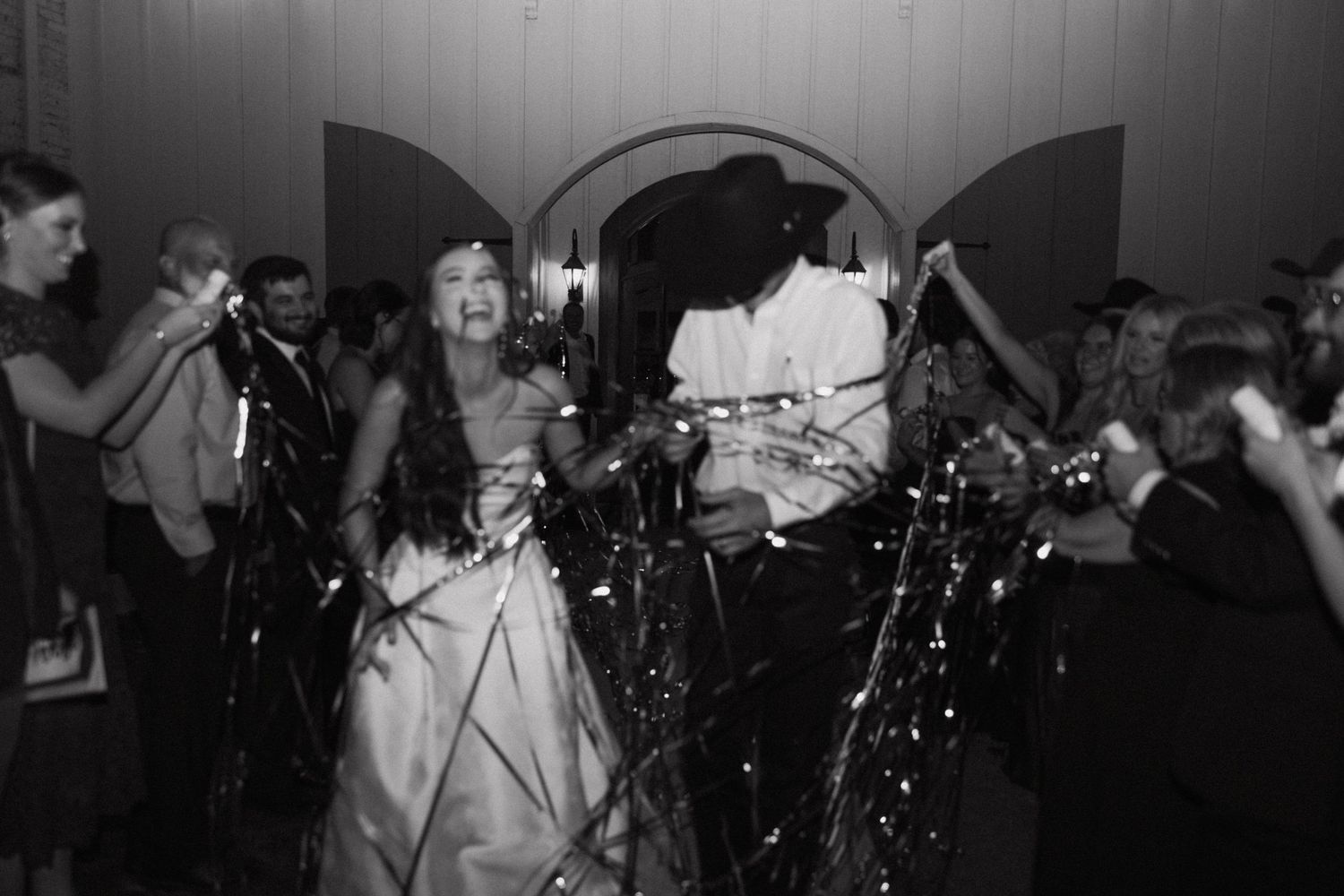 A black and white photo of a wedding celebration with guests throwing confetti and streamers in a dimly lit venue.