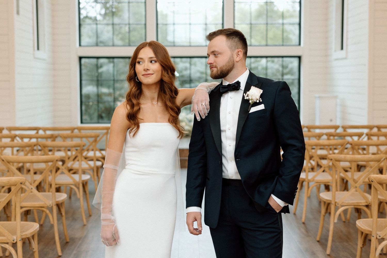 A couple in wedding attire stands together in a bright chapel with wooden chairs and large windows.